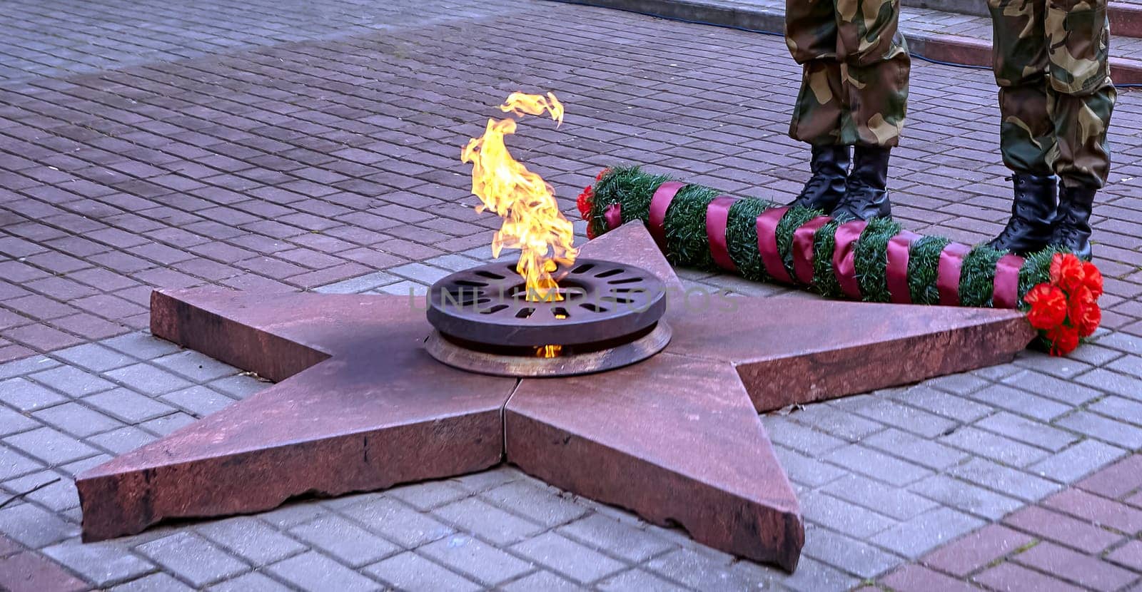 A soldier in camouflage uniform and boots stands at a monument with an eternal flame. A wreath and flowers honor the fallen. The soldier symbolizes dedication and respect.