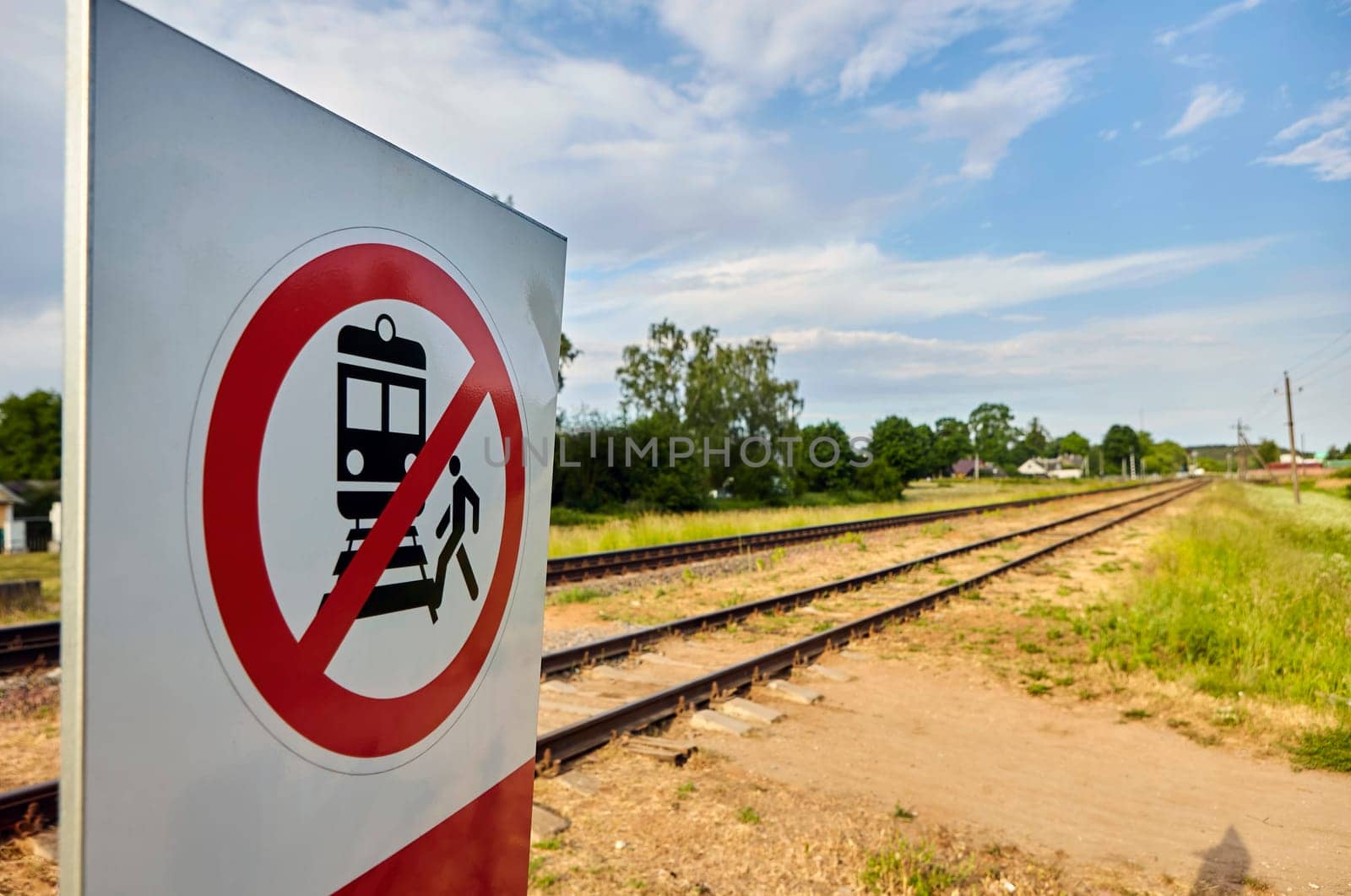 A concrete railway track under a sunny sky with a vivid prohibition sign displaying a train and a person, emphasizing no crossing. The scene captures rail safety on a lovely day.