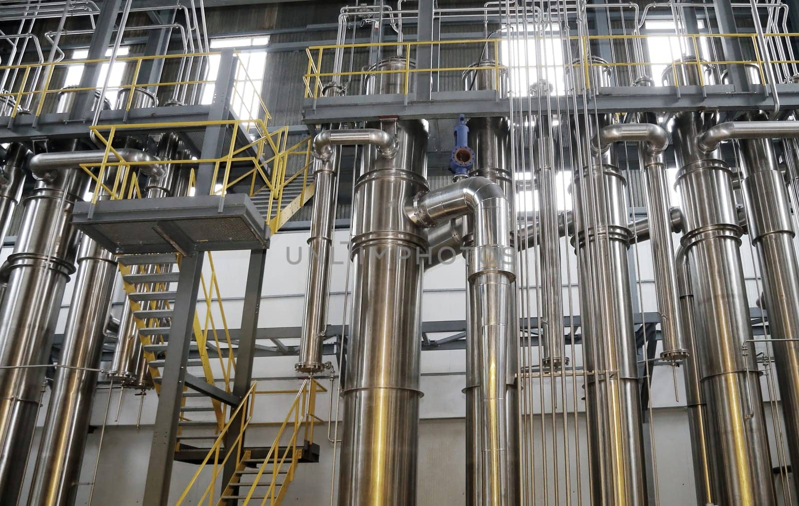 A chemical plant features a complex network of stainless steel pipes and glass vessels, creating an intricate system of interconnected straight and curved components in various sizes and shapes.