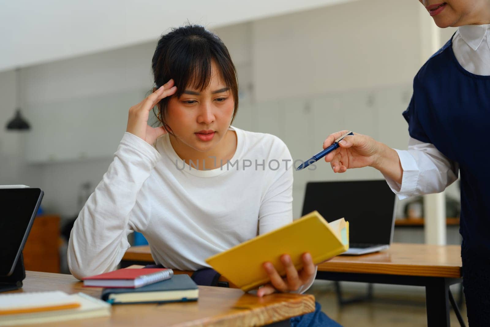 Mature lecturer helping female student struggling with schoolwork in a classroom by prathanchorruangsak