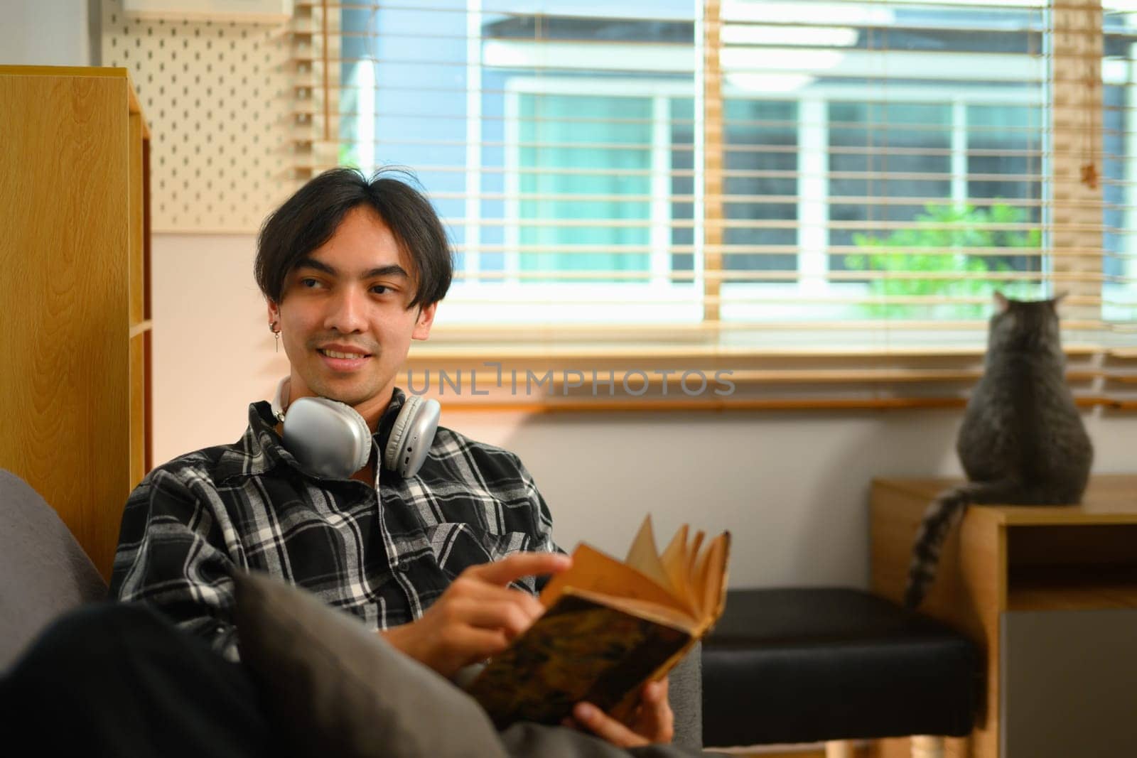 Pleased young man holding book sitting on couch and looking away.