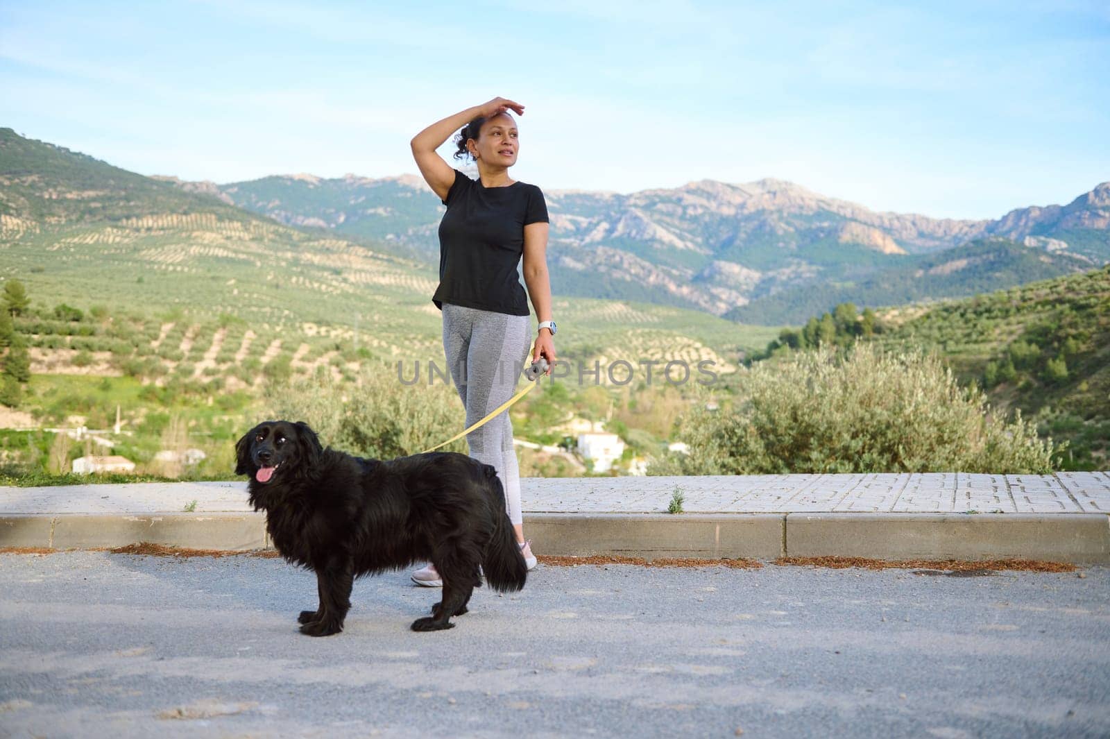 Beautiful woman in sportswear, looking way, walking her dog on leash in the nature, enjoying walk with her pedigree purebred black cocker spaniel dog in the mountains nature outdoors by artgf