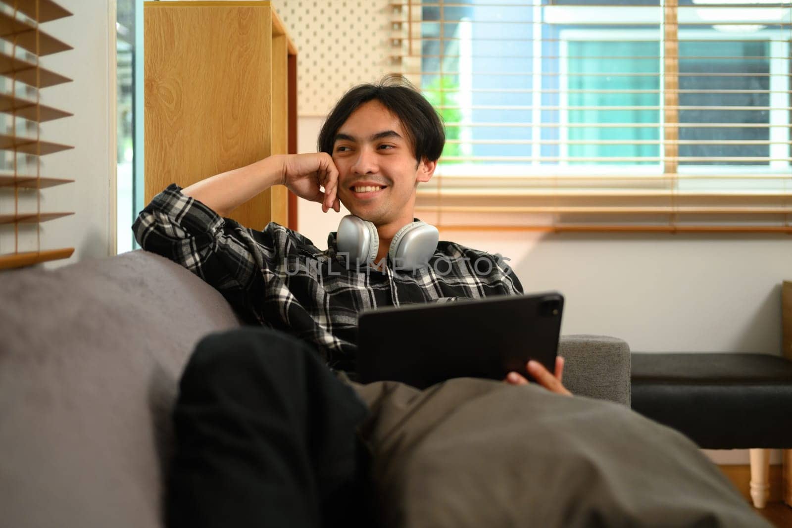 Happy young Asian man looking away while sitting with digital tablet on couch at home by prathanchorruangsak