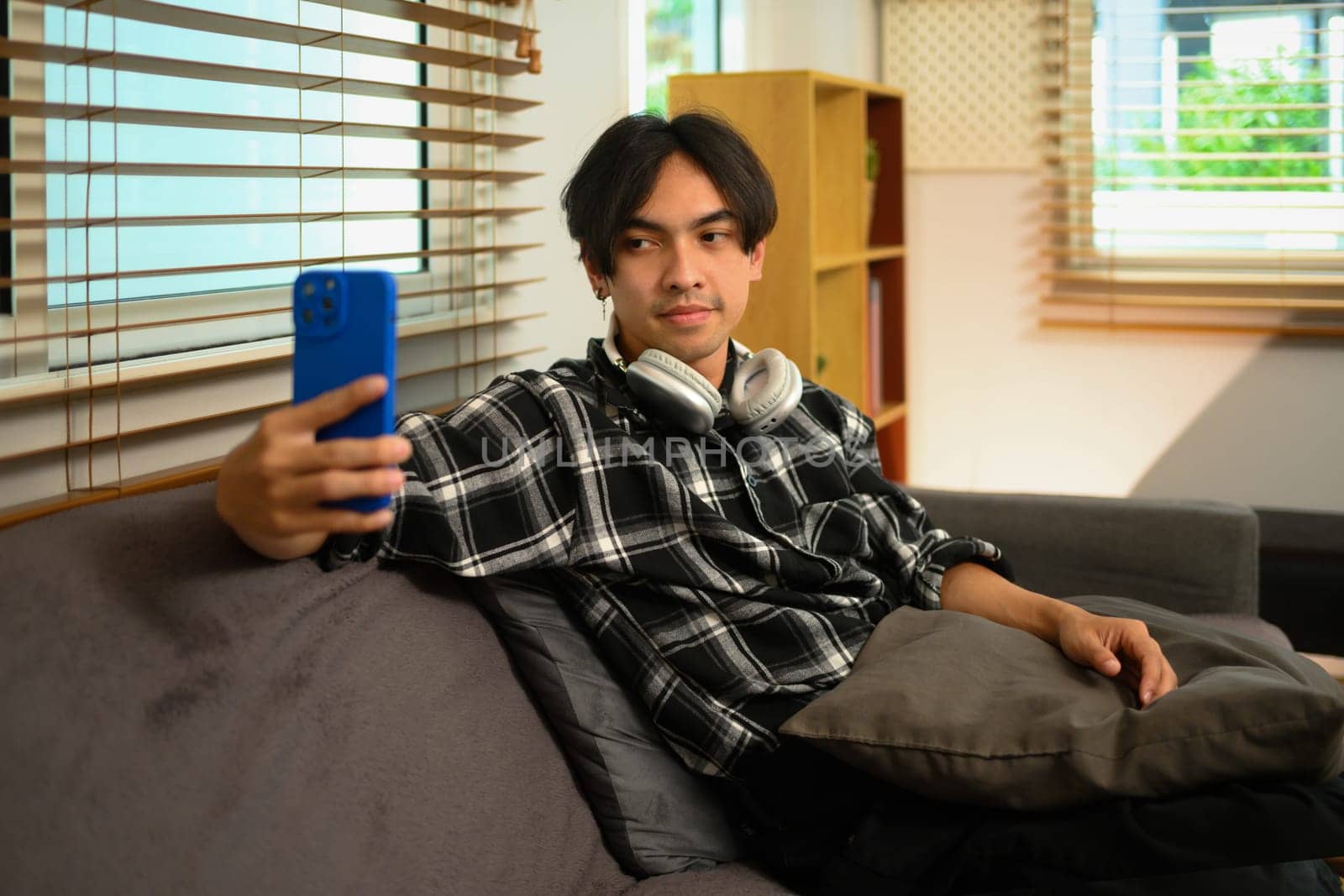Portrait of young smiling man reading message on mobile phone, relaxing on couch at home.