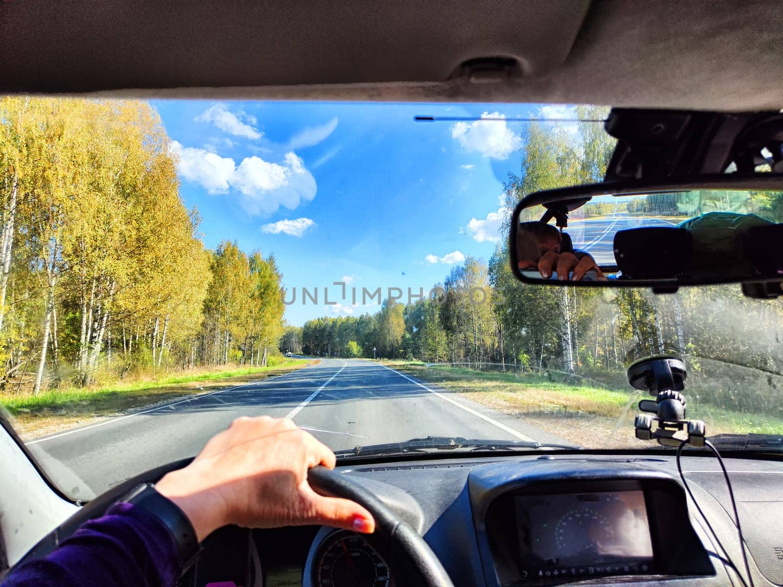 view from a car windshield of natural landscape with road, green trees and blue sky in summer or spring time. Hand of woman on steering wheel. Female traveler driving alone on trip or journey
