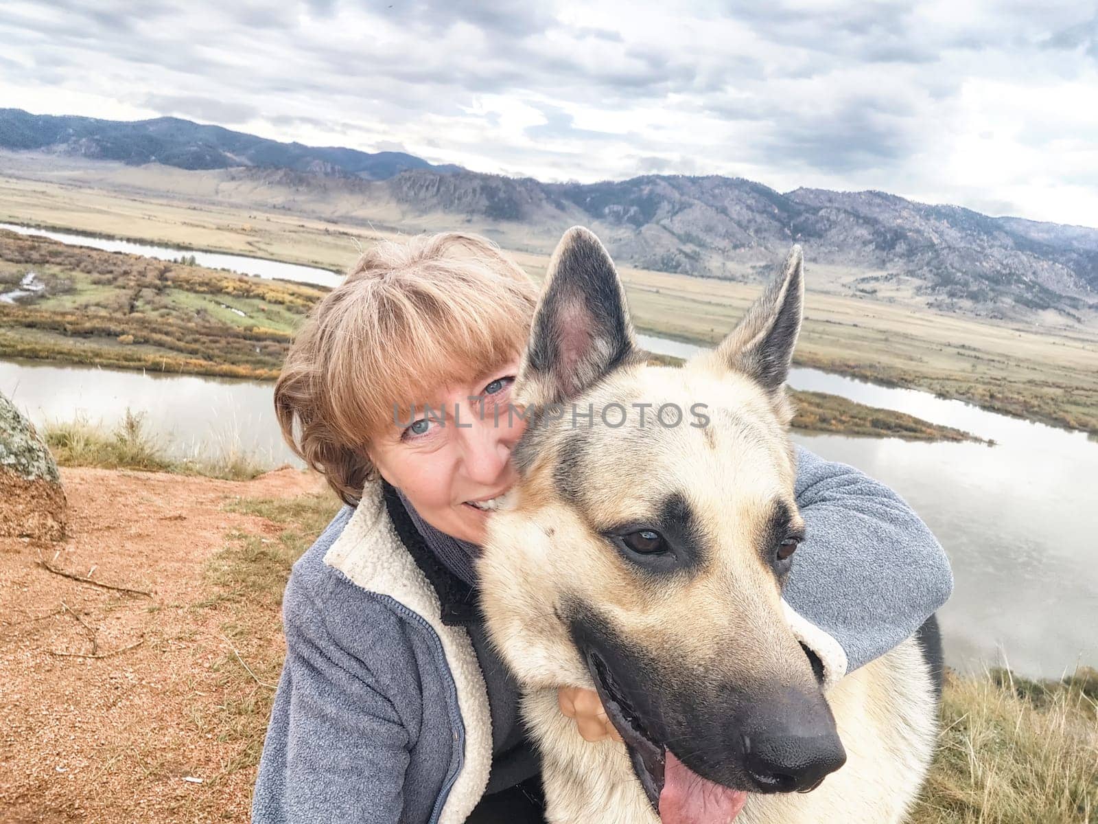 Adult girl with shepherd dog taking selfie near water of lake or river in mountain. Middle aged woman and big pet on nature. Friendship, love and fun