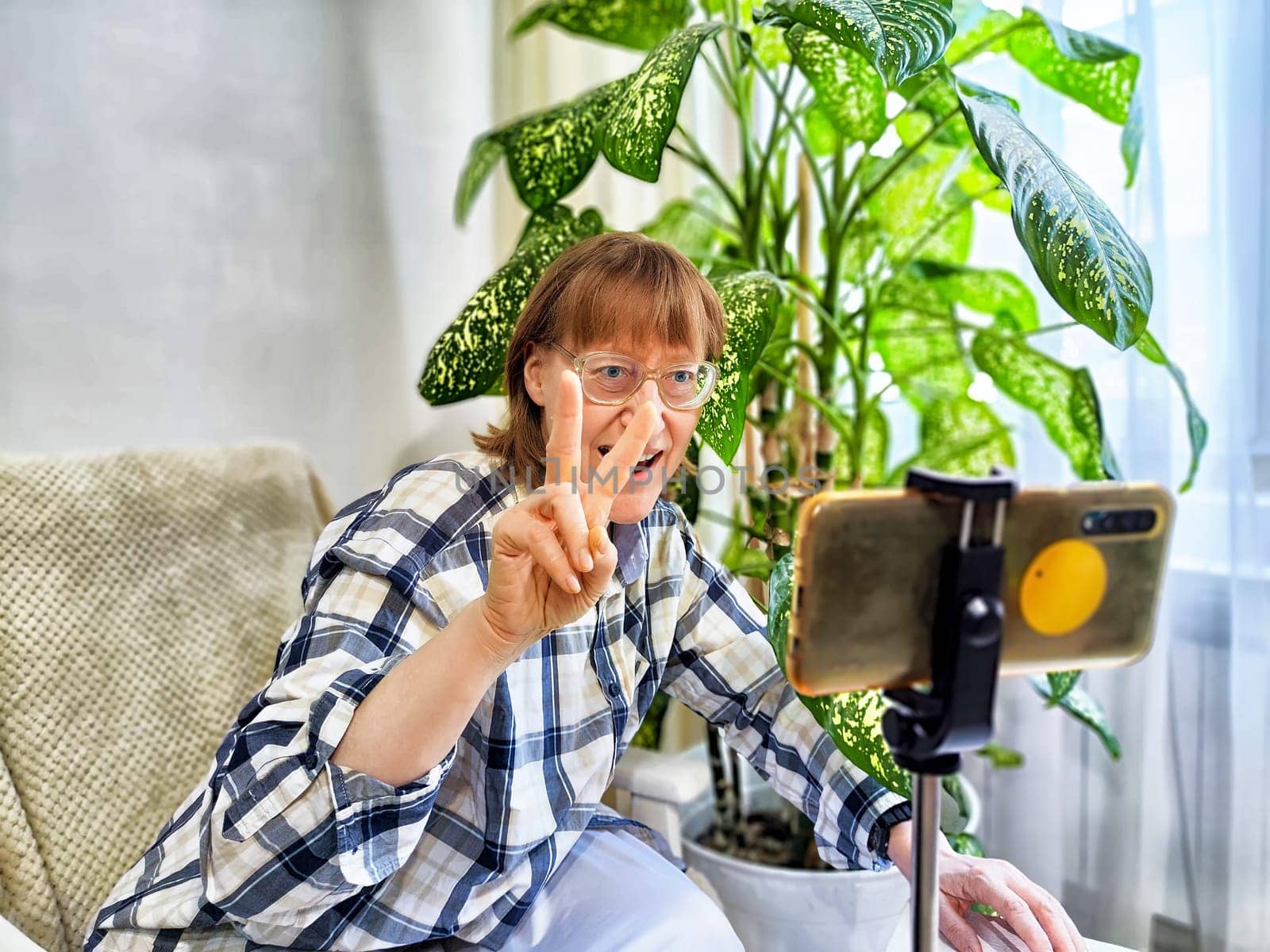 Smiling woman in glasses using a smartphone on a tripod for a video chat. A middle-aged woman posing and taking selfie by keleny