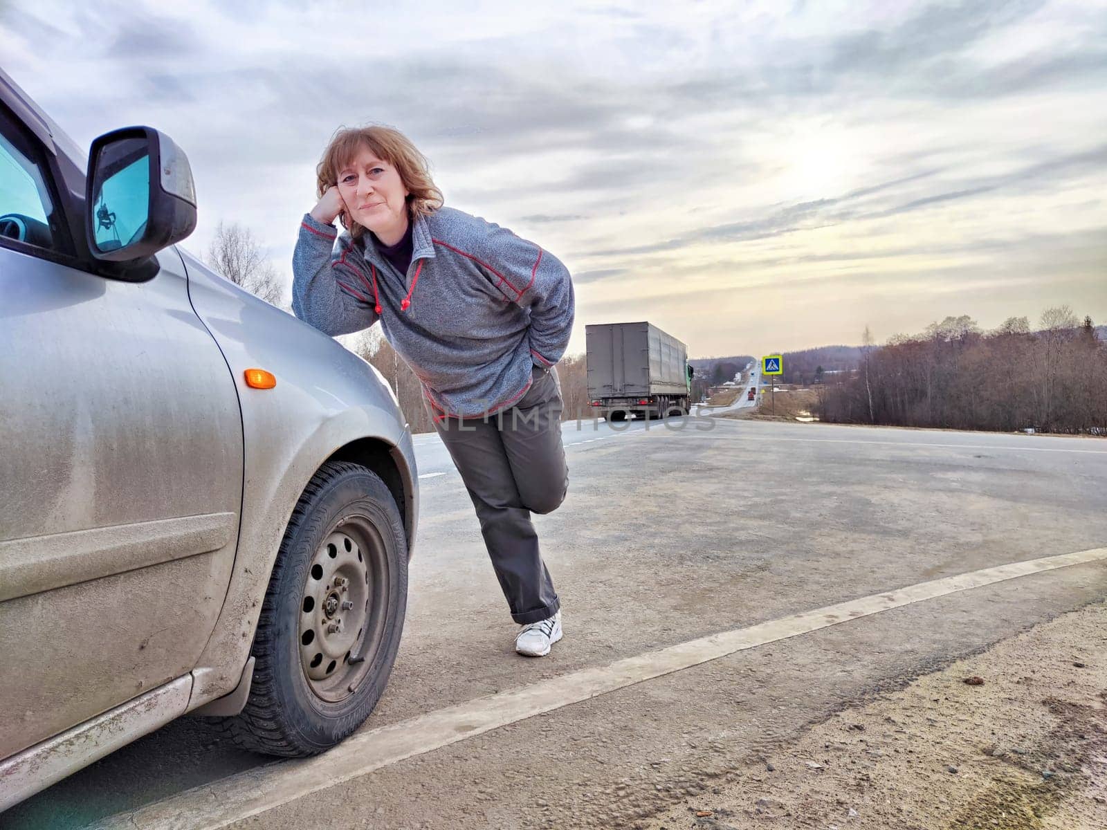 Girl Resting on a Road Trip at Dusk by Her Car. A traveler takes a break, leaning on her parked car on an open road, with the sky hinting at dusk by keleny