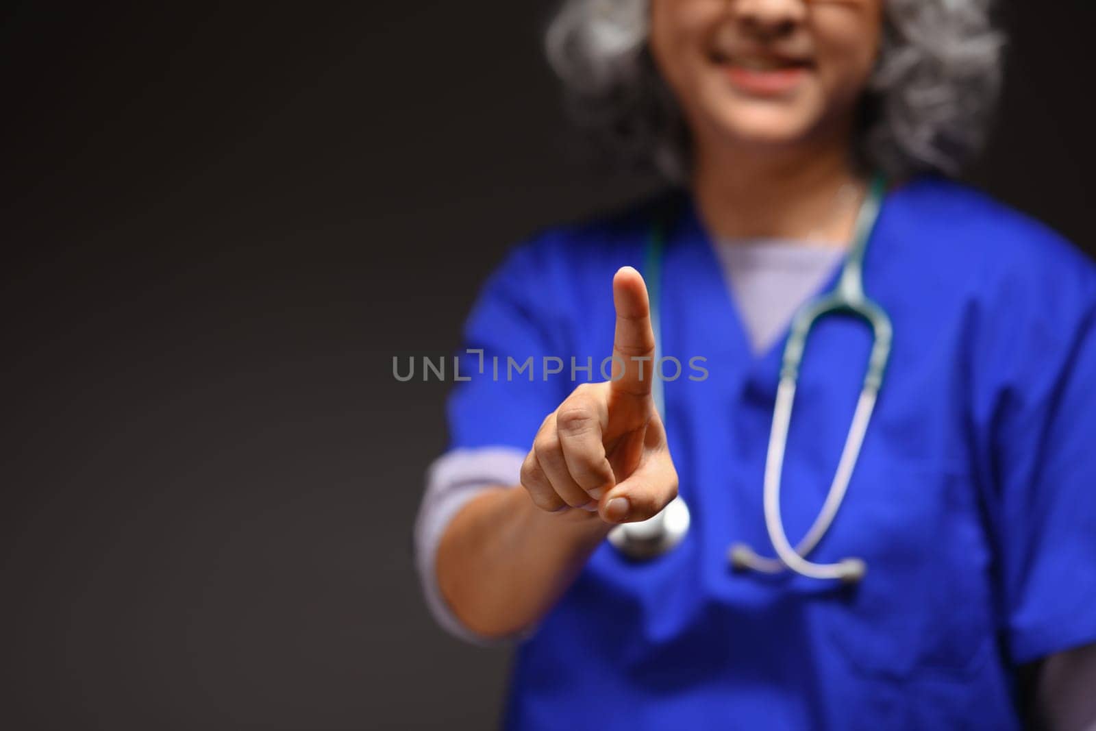 Doctor with stethoscope standing against black background and pointing towards the camera.