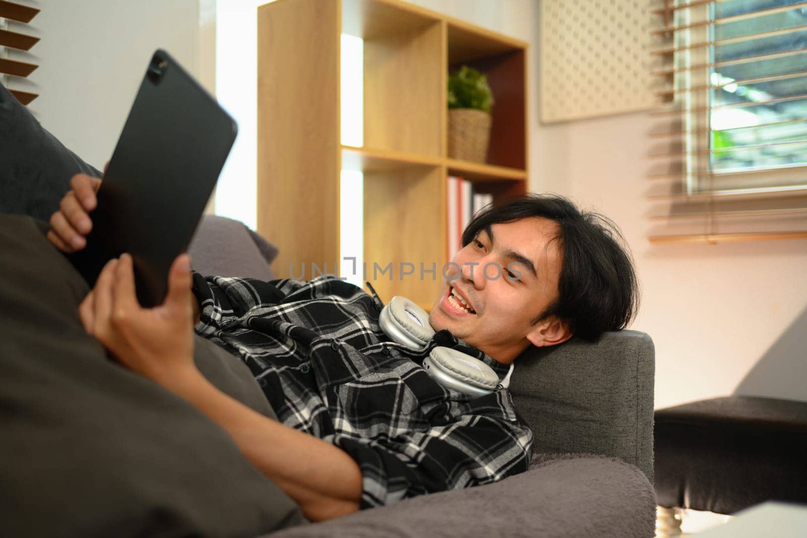 Cheerful young man lying on couch and watching movie on digital tablet at home.