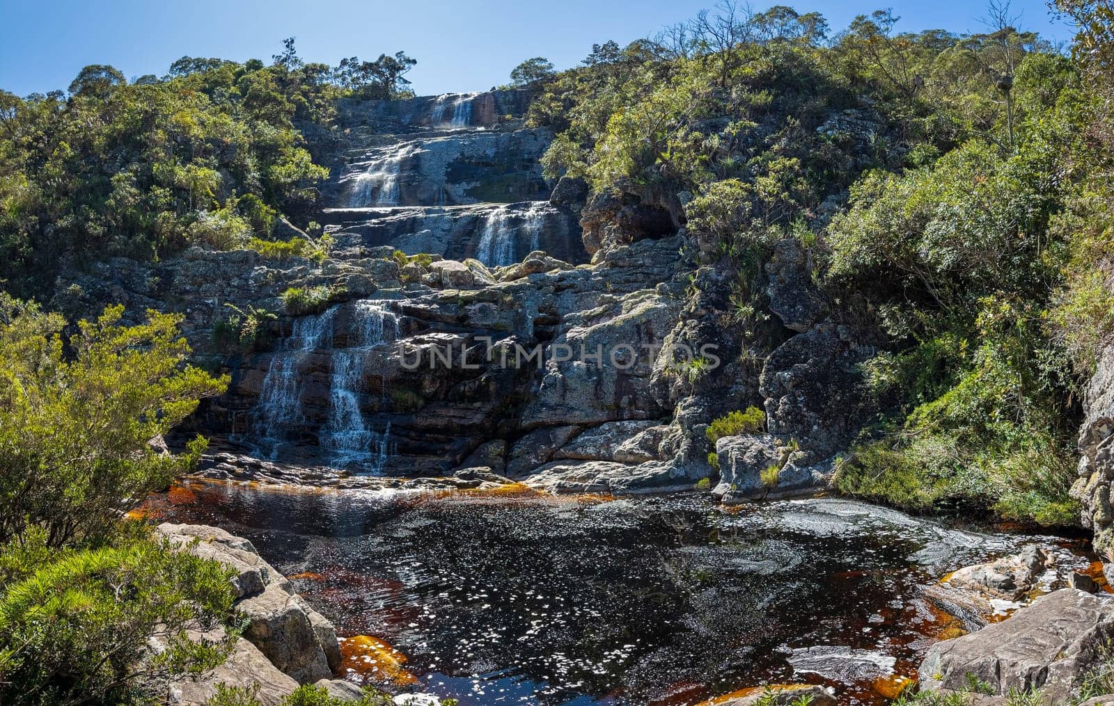 A picturesque, layered waterfall set amidst lush greenery and a bright blue sky.