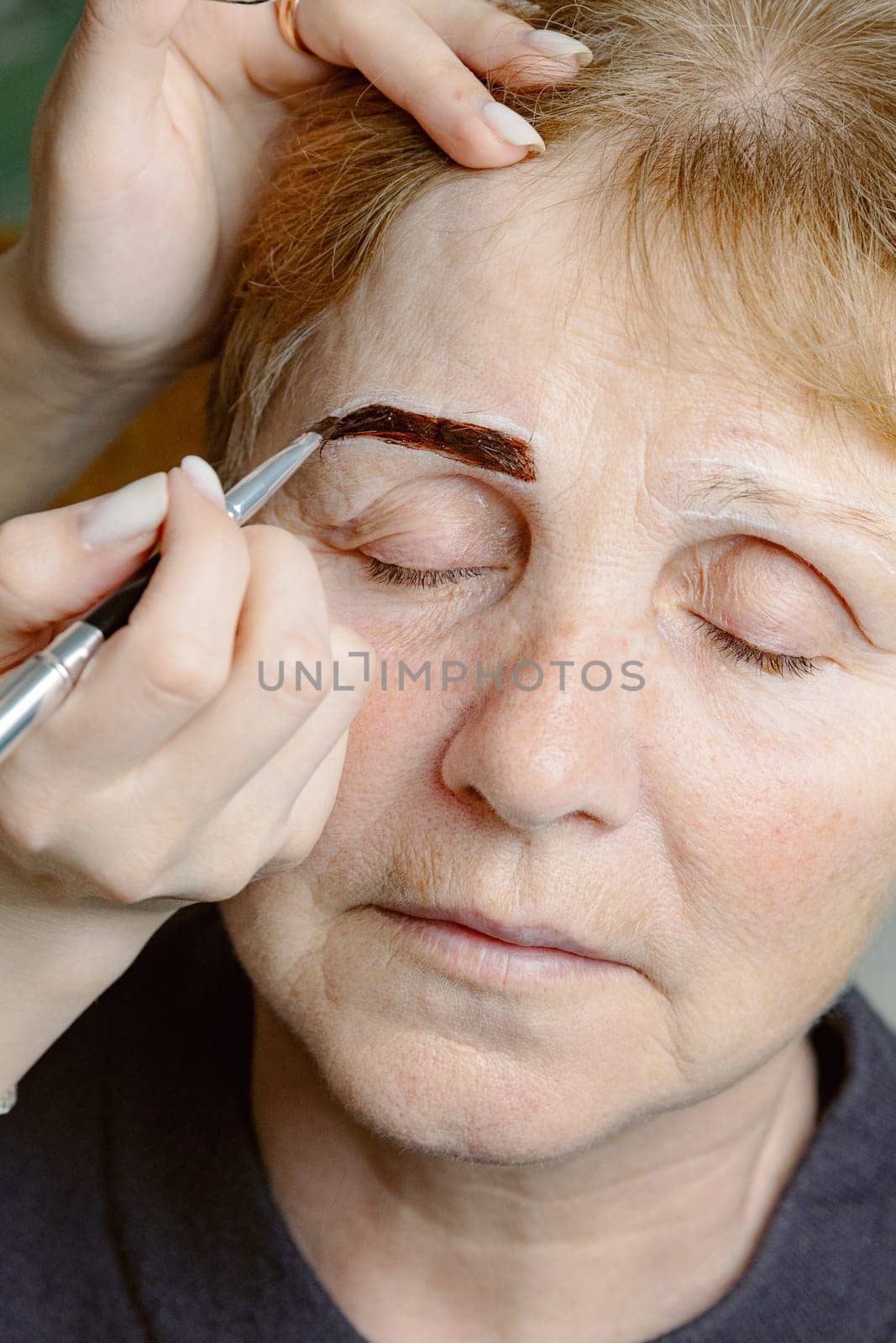 One young Caucasian young unrecognizable girl cosmetologist carefully applies dark brown paint with a brush on the right eyebrow of an elderly woman with closed eyes, sitting on a white background in a home beauty salon, top view very close up.