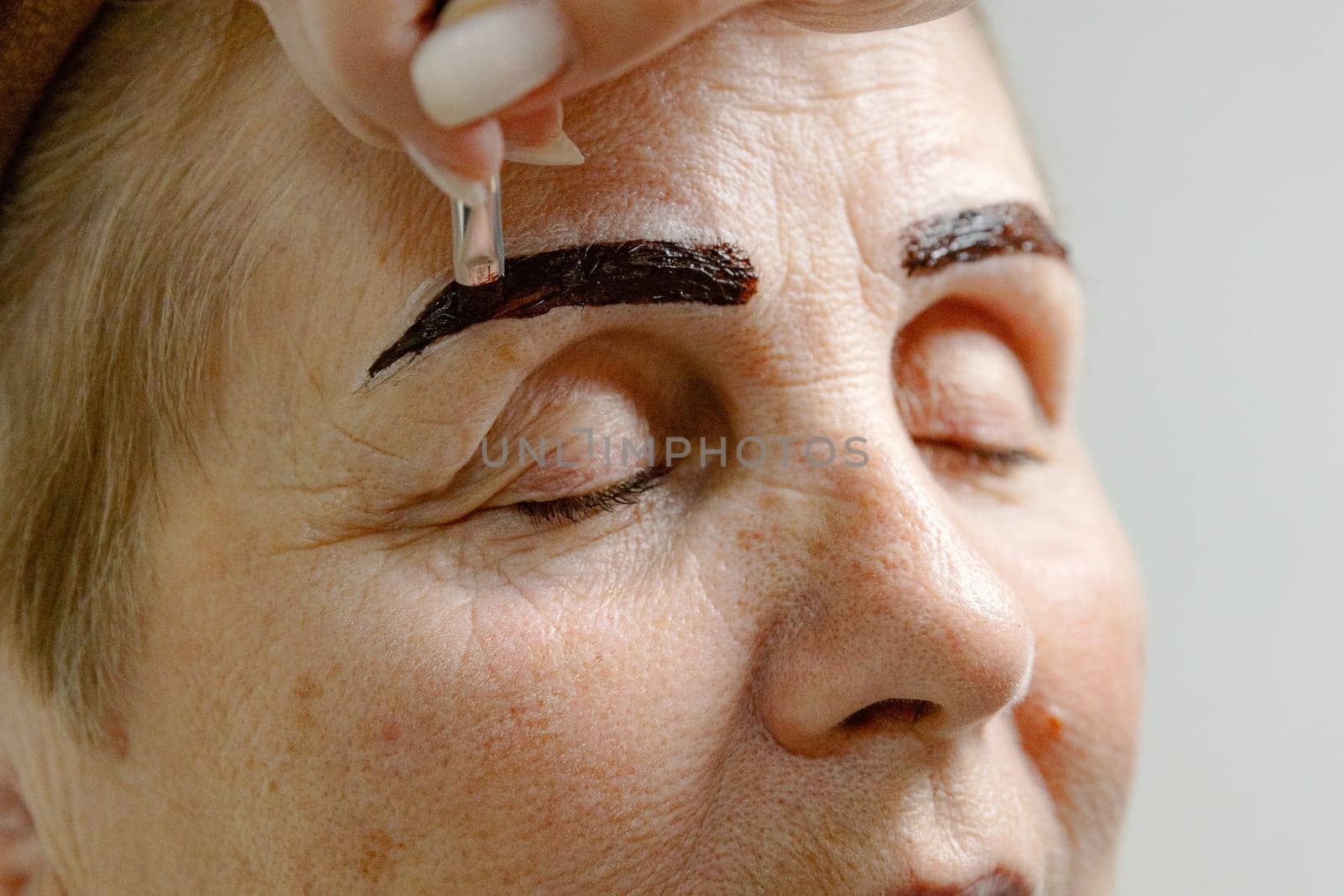 One young Caucasian unrecognizable young girl cosmetologist carefully applies dark brown paint with a brush on the right eyebrow of an elderly woman with closed eyes, sitting on a white background in a home beauty salon, side view very close up.