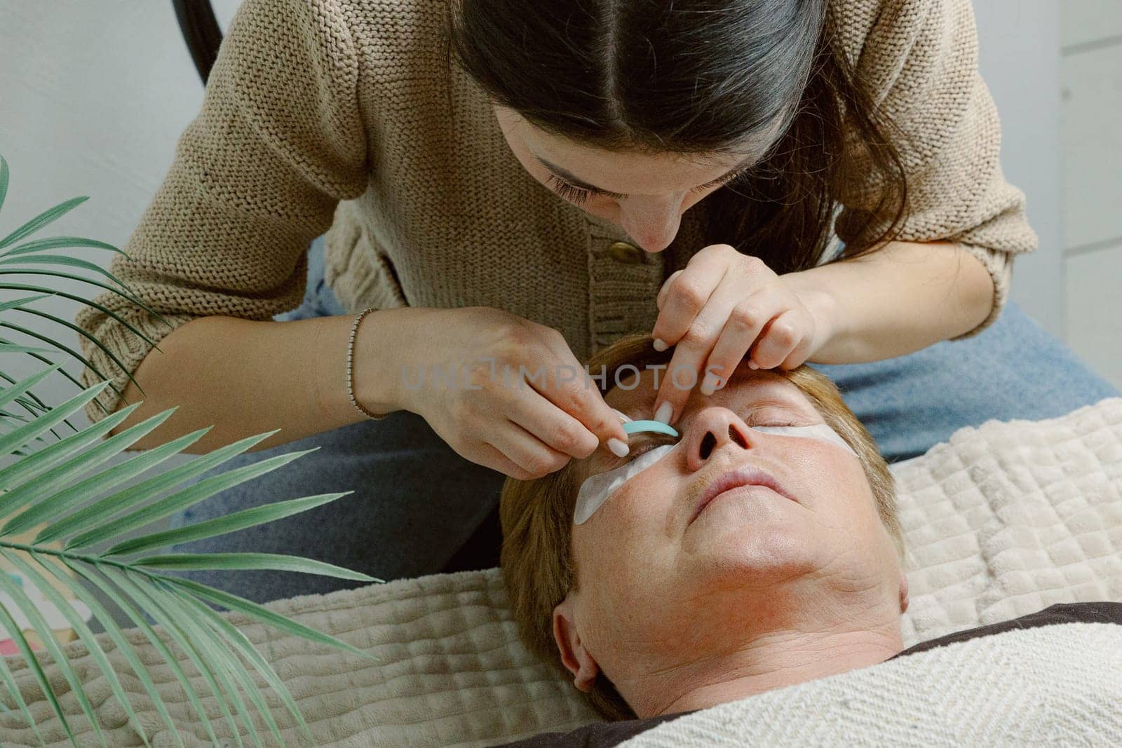 One Caucasian young beautiful brunette girl cosmetologist sticking a blue silicone eyelash stand for the right eye of an elderly client who is lying on a cosmetology bed in a home beauty salon, top side close-up view.