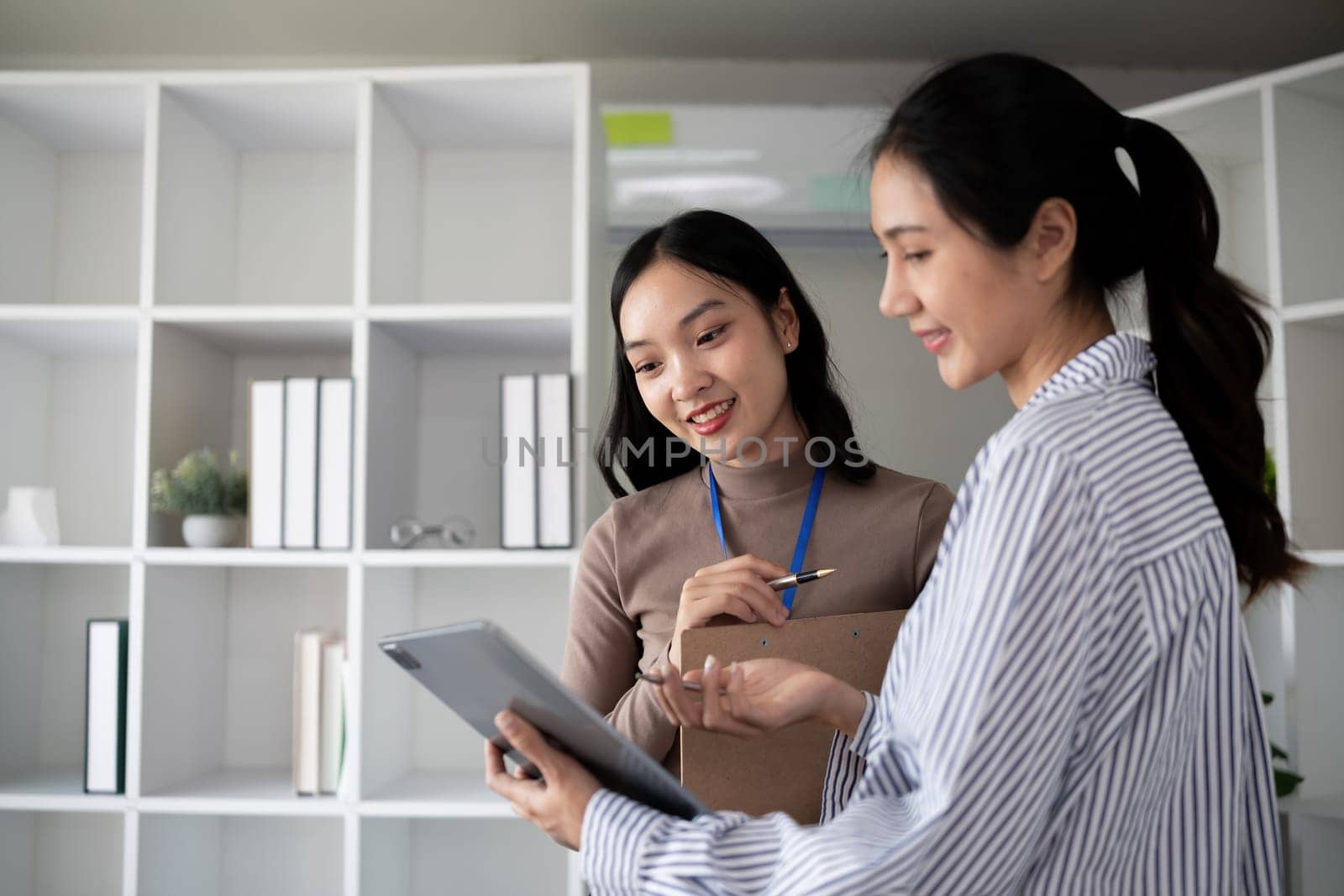 Two Asian businesswomen discussing work using a tablet in a modern office. Concept of teamwork and professional collaboration.