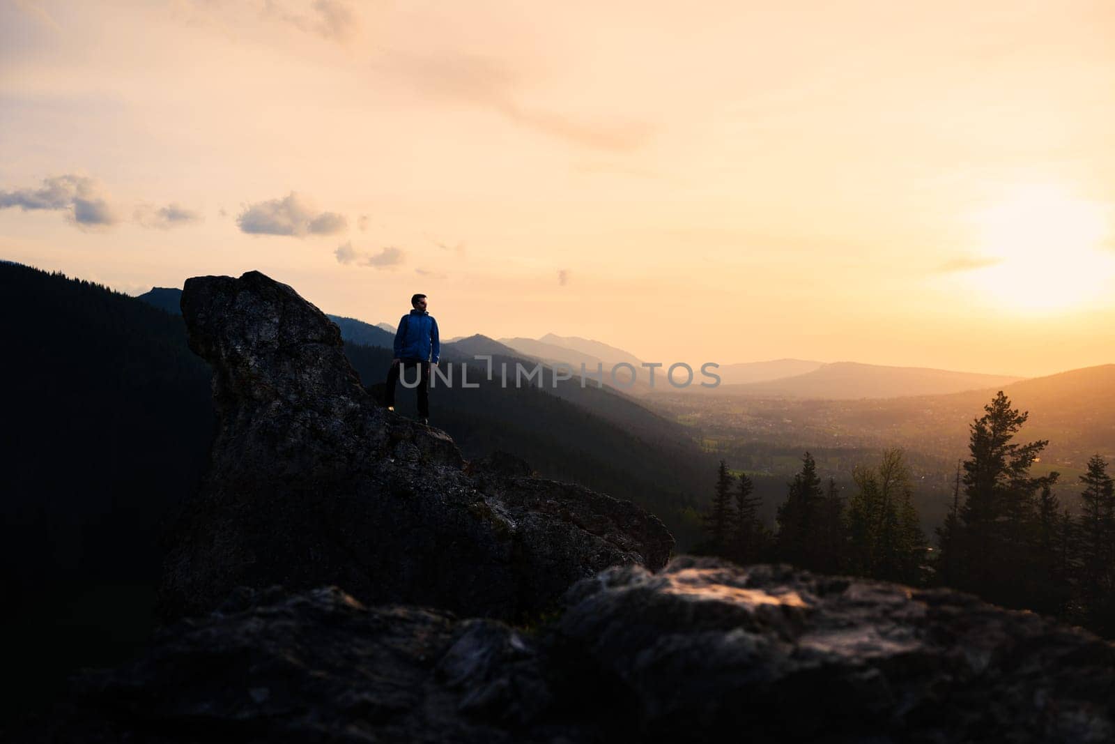 At dusk, a person stands on a mountain peak, surrounded by the natural landscape. The sky is painted with vibrant colors of the sunset