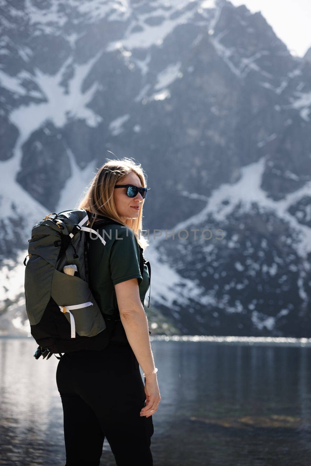 A traveler girl with a backpack enjoys the stunning view of a mountain range and a lake, with snow-capped peaks in the background.