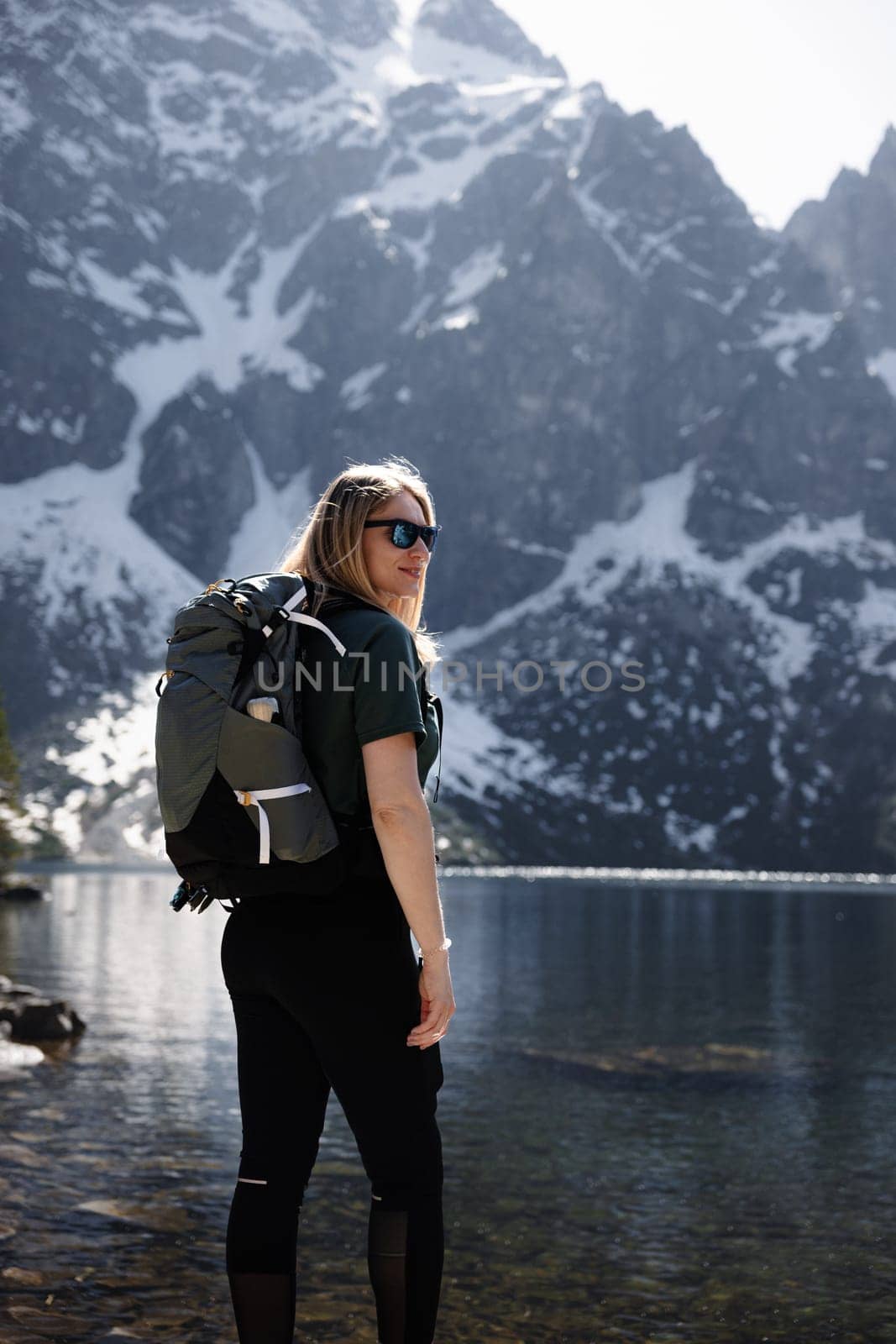 Blonde woman with backpack by lake, mountains and snow in backdrop by Kustov
