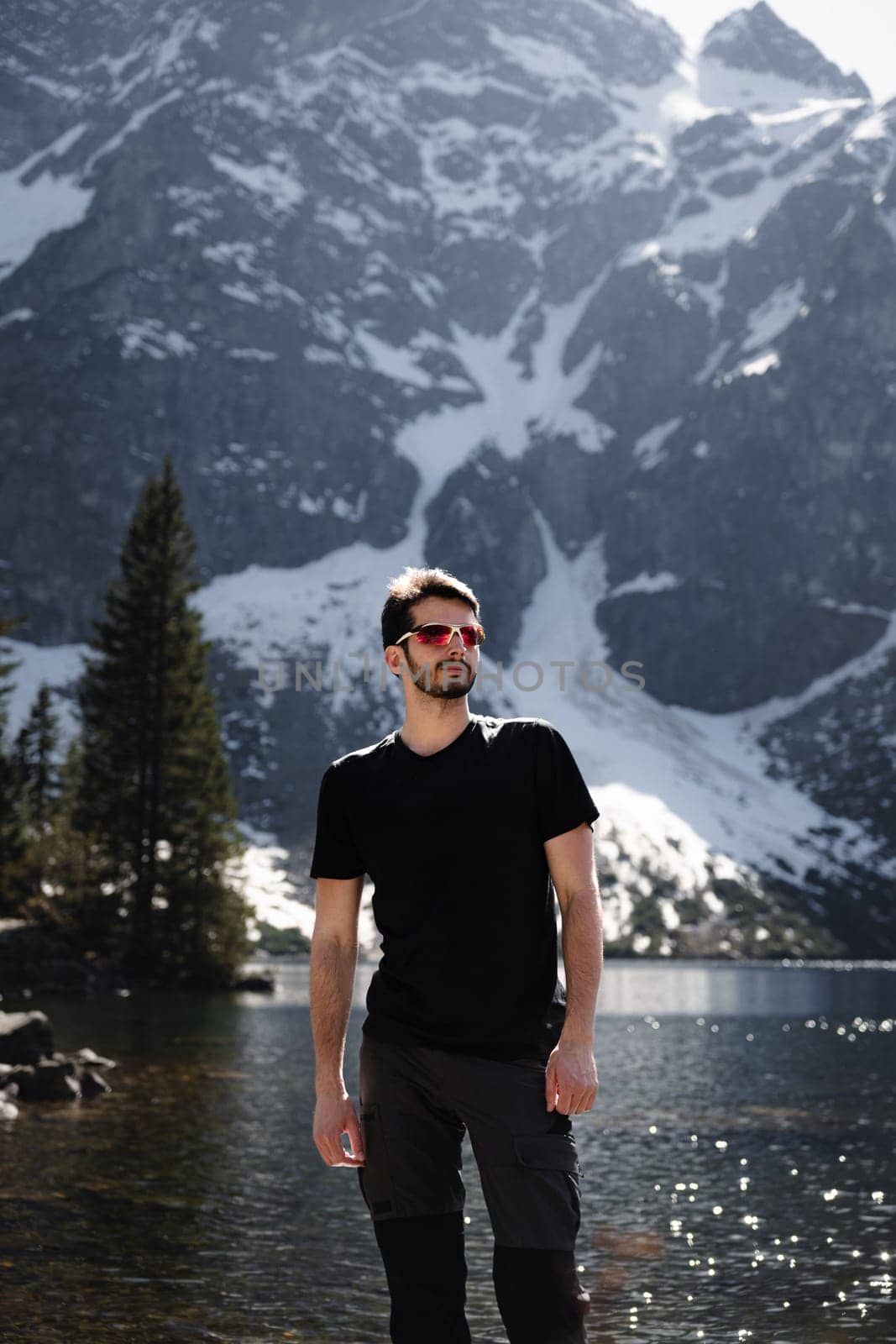 A man wearing a black shirt and sunglasses is standing by a lake in front of a majestic mountain, enjoying the beautiful natural landscape