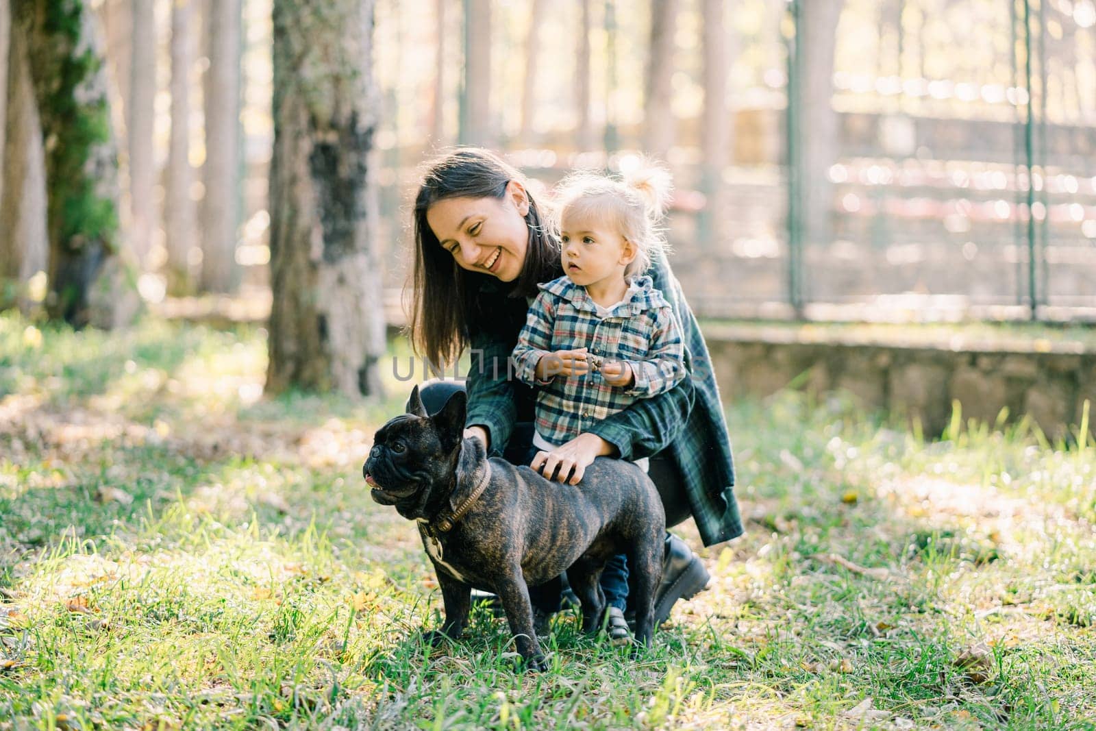 Little daughter stands next to smiling mother stroking french bulldog in forest. High quality photo
