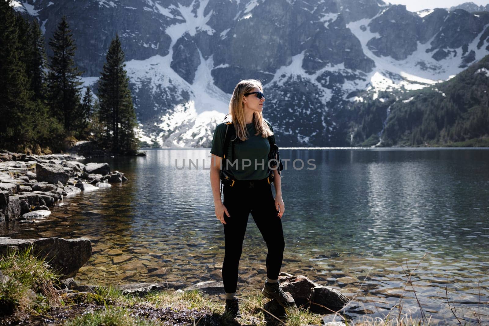 Blonde woman with backpack standing at full height by lake, mountains and snow in backdrop by Kustov
