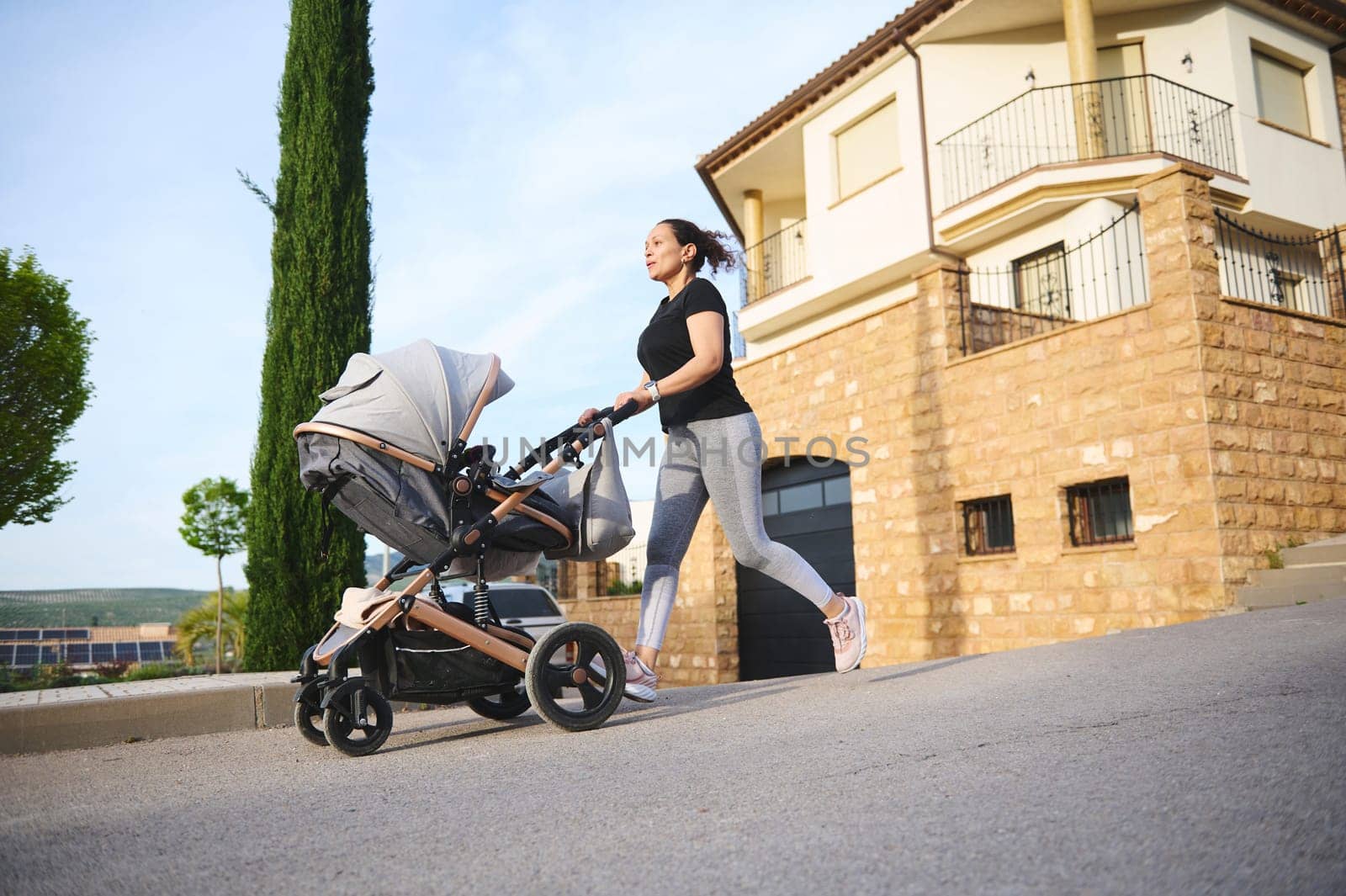 Happy woman performing morning jog and pushing a baby pram, enjoying her active lifestyle during maternity leave. People. Motherhood and babyhood concept by artgf