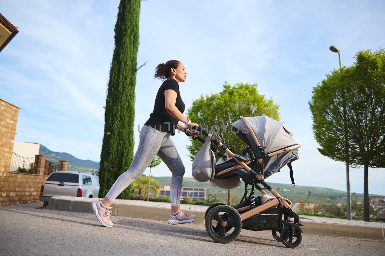 Full length shot of happy woman performing morning jog, pushing a baby pram running on the city street and enjoying her active lifestyle during maternity leave. People. Motherhood and babyhood concept