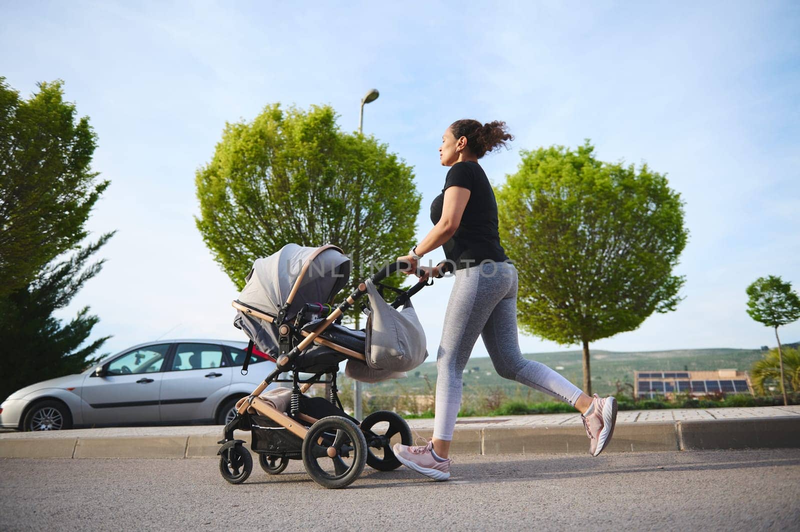 Full length photography in motion of an active woman exercising outdoors, running morning jog while pushing her baby pram. People. Active and healthy lifestyle. Maternity and fitness with baby concept