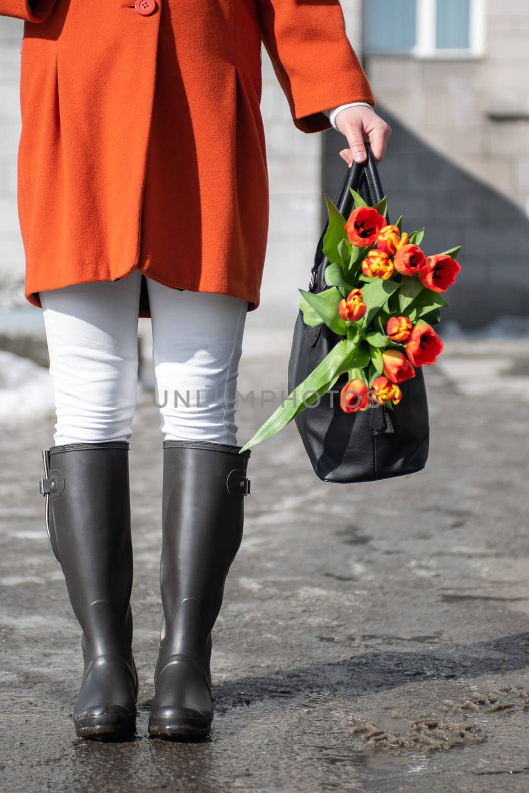 A beautiful young woman in a orange coat and rubber boots walking down the street with a bouquet of tulips in spring city