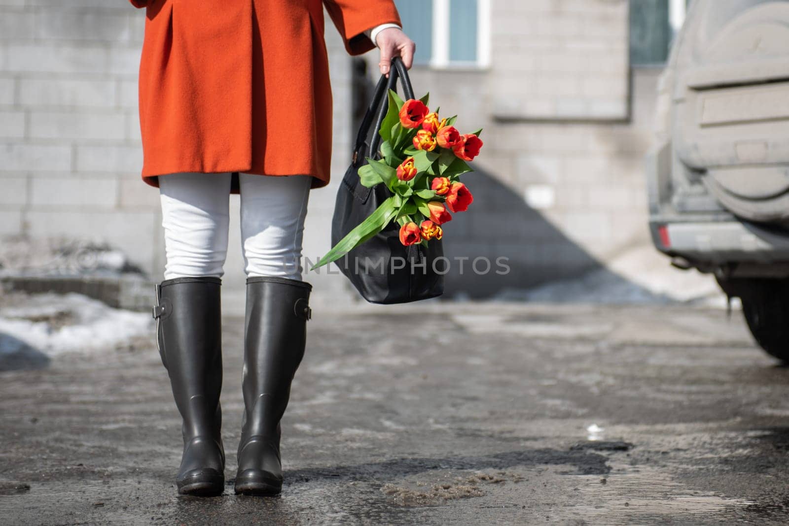 A beautiful young woman in a orange coat and rubber boots walking down the street with a bouquet of tulips by rusak