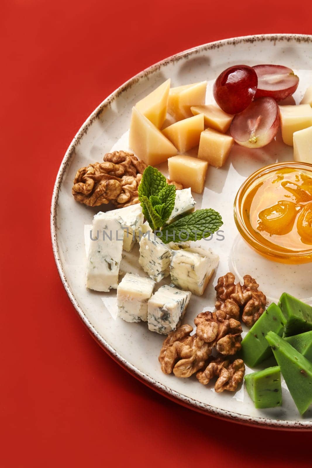 Artfully served artisan cheese board featuring blue and green pesto cheeses, purple grapes, walnuts, and sweet honey on ceramic dish with rich red backdrop. Popular Italian style snack