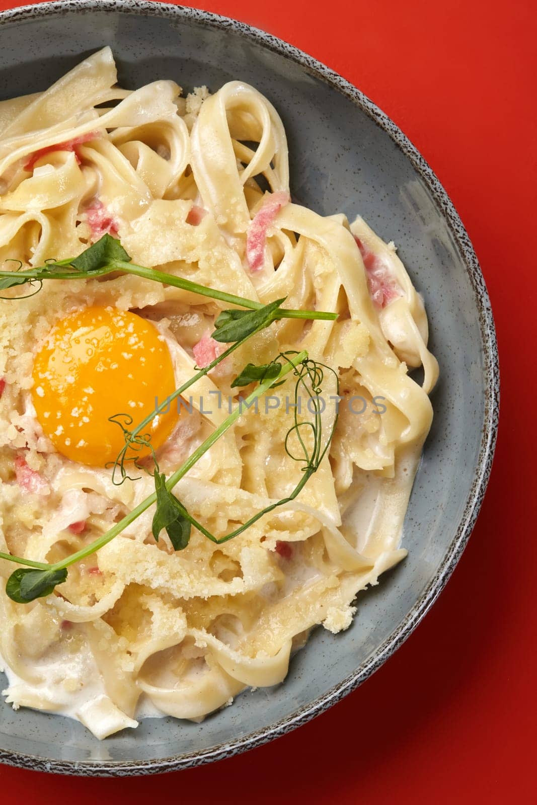Detailed view of classic fettuccine carbonara with chunks of pancetta and vibrant egg yolk sprinkled with grated parmesan and garnished with pea shoots, served in gray ceramic plate on red backdrop