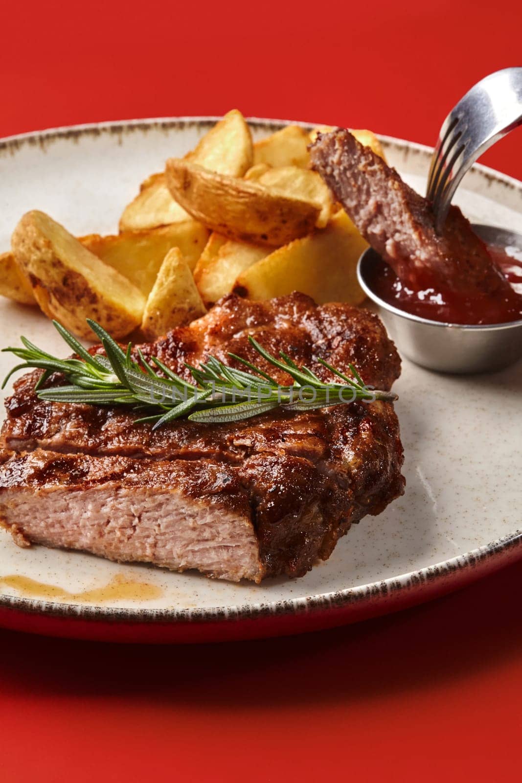 Succulent sliced medium rare pork steak served with crispy baked potato wedges and tangy barbecue sauce, presented elegantly on textured plate against vibrant red backdrop