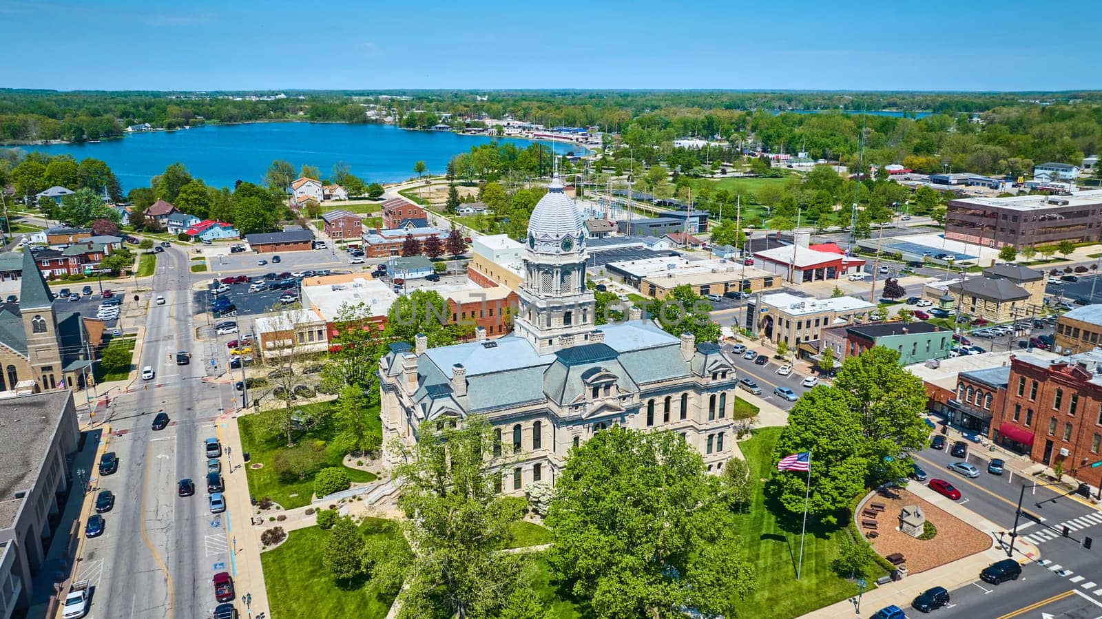 Aerial view of Warsaw, Indiana, featuring the historic Kosciusko County Courthouse surrounded by vibrant town life and scenic lake.