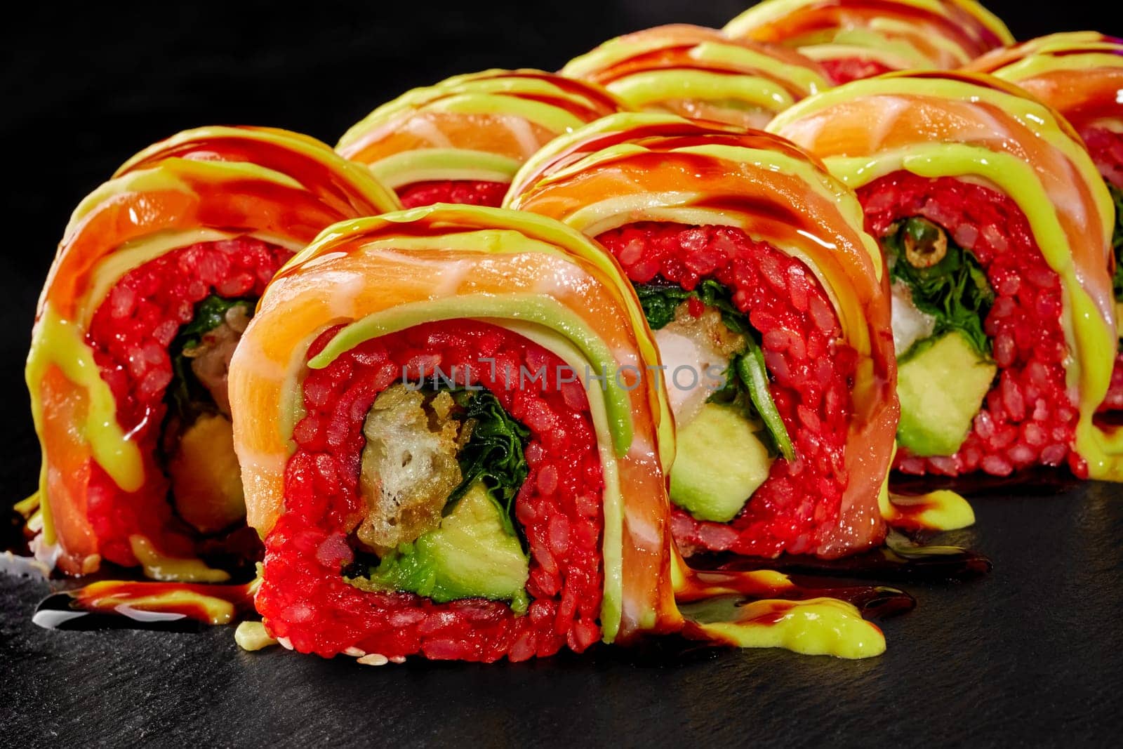 Macro shot of decadent sushi rolls with shrimp tempura, covered in slices of salmon and drizzled with savory sauces, served on black stone surface for luxurious Japanese style dining experience