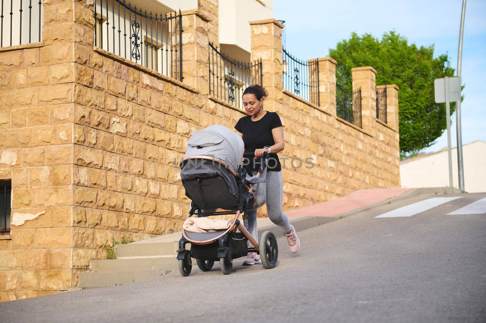A smiling young woman in black t-shirt and gray sports leggings, pushing baby buggy while exercising outdoors. People. Maternity and infancy. Fitness with baby. Active and healthy lifestyle concept
