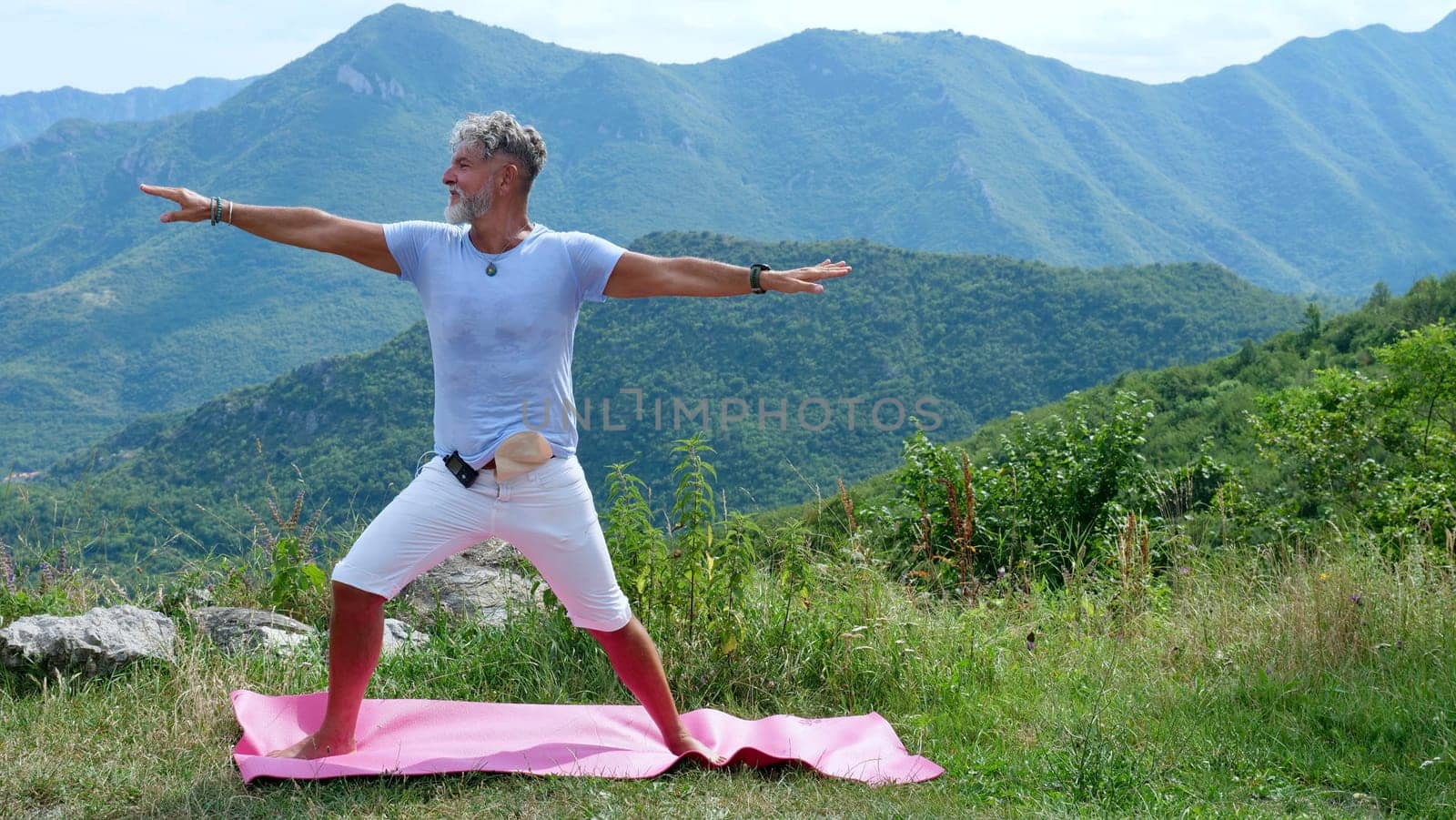 A sick old gray-haired man with diabetes and colon cancer with a colostomy practices yoga in the mountains. Stands in the pose of a warrior