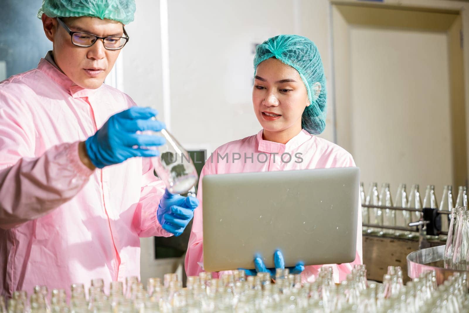 Two engineers oversee quality control checking product bottles on a beverage factory's conveyor belt by Sorapop