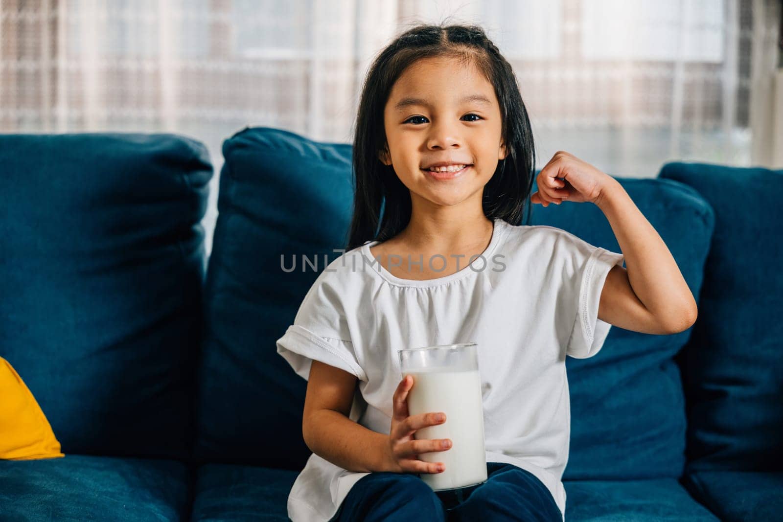 Close-up of happy Asian girl holding glass of milk her delighted face showcasing joy of a nutritious drink on the cozy sofa at home. A heartwarming portrayal of daily health care and childhood bliss.