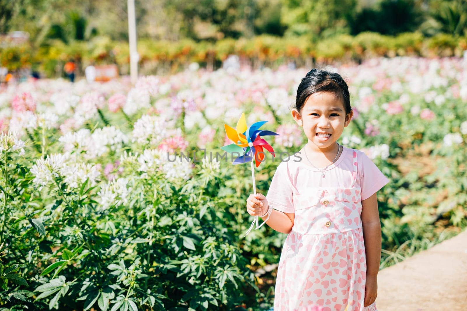 Portrait of a joyful girl in a flower garden holding a toy pinwheel. The lively spring season evokes happiness and the spinning pinwheel embodies childhood joy and freedom in nature's sunny beauty.