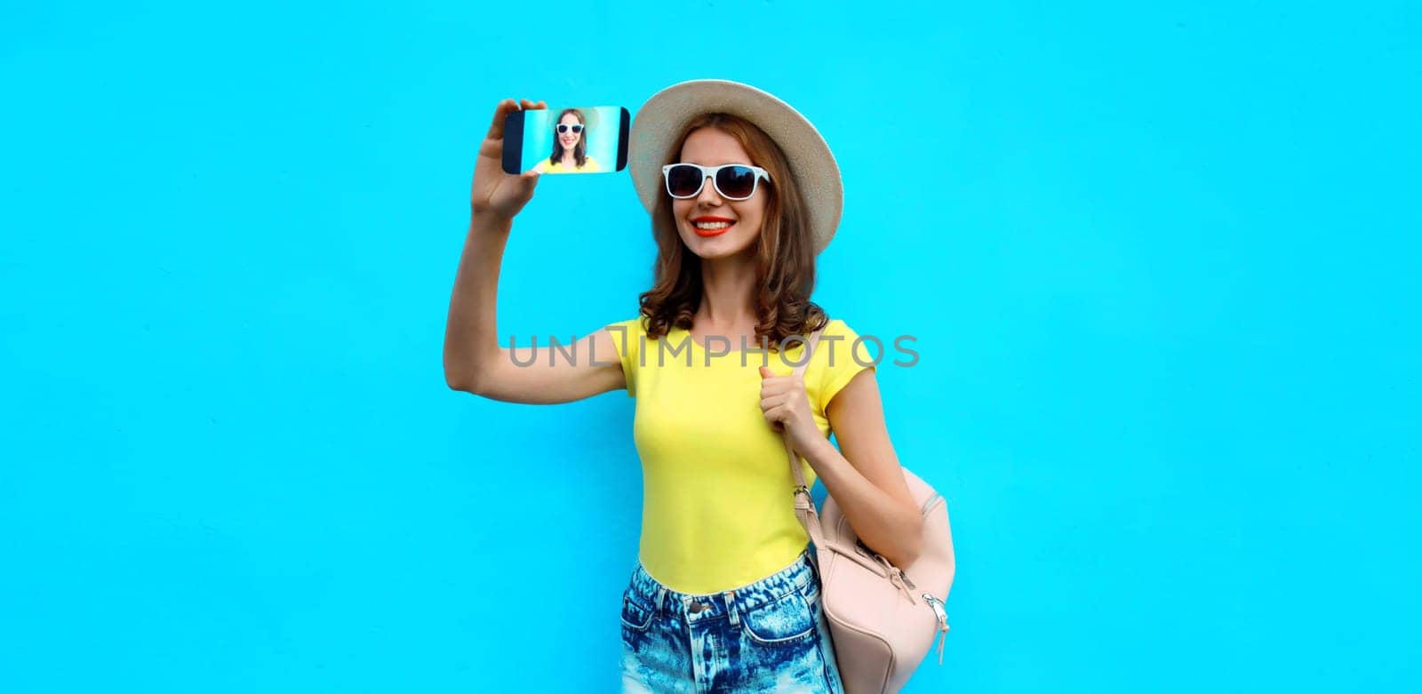 Portrait of happy smiling young woman taking selfie with smartphone wearing a summer straw hat with backpack on blue background