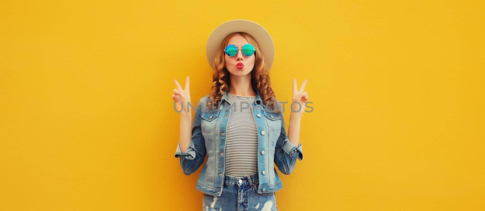 Portrait of beautiful stylish young woman blowing kiss in summer hat, sunglasses, denim jacket posing on yellow studio background