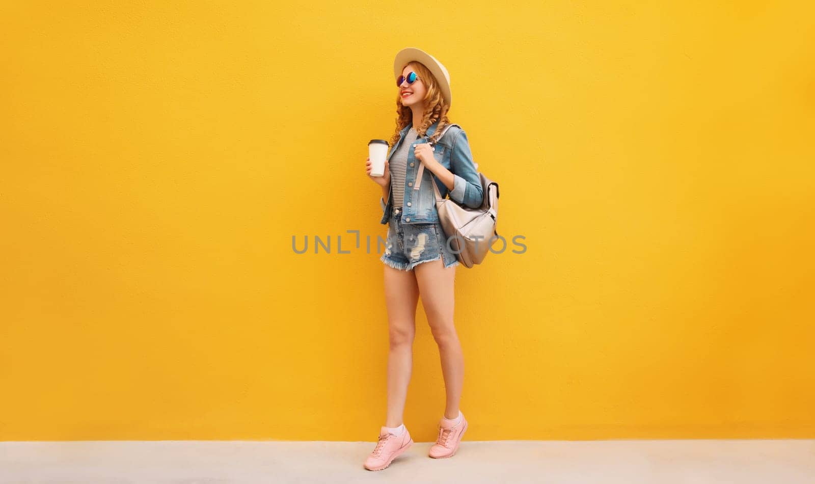 Beautiful stylish happy smiling young woman with cup of coffee wearing summer hat, backpack, jean jacket on yellow background