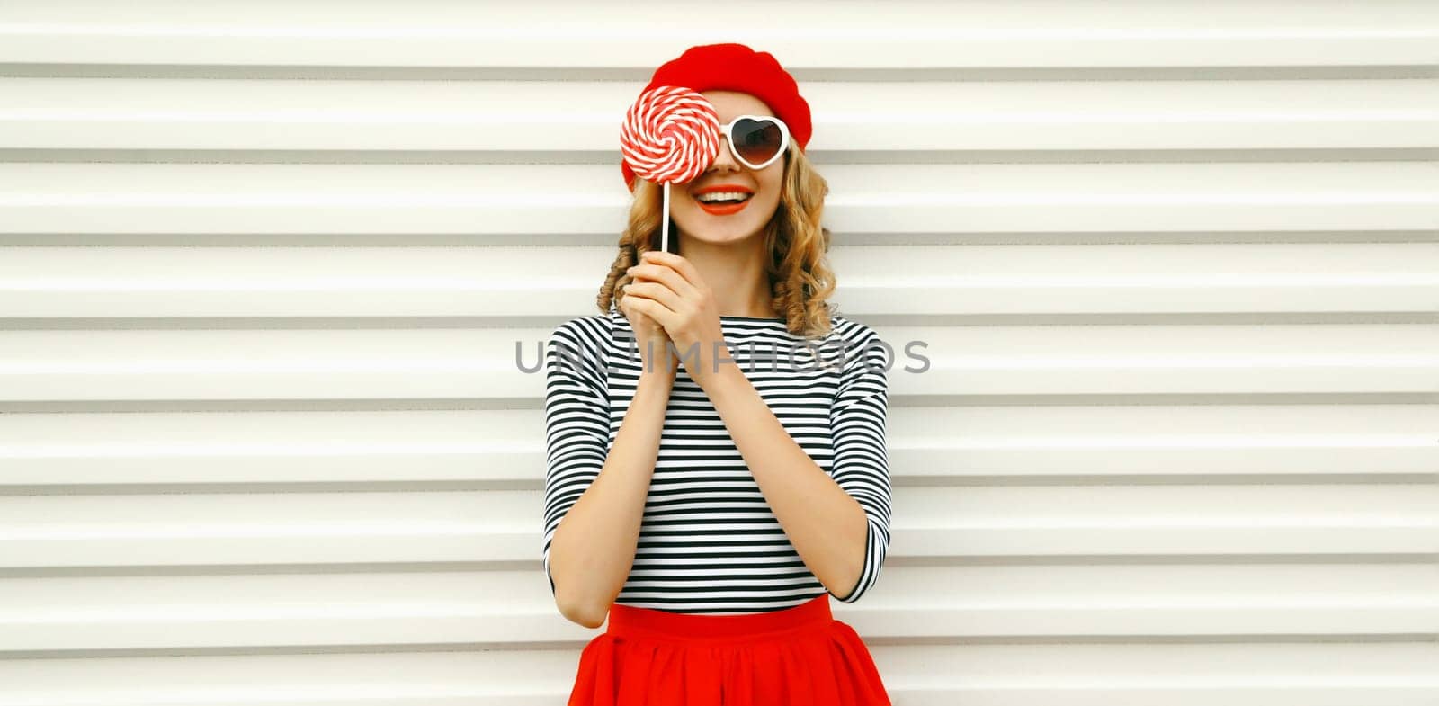 Portrait of happy cheerful smiling young woman holding colorful lollipop wearing white sunglasses, red beret hat on white background
