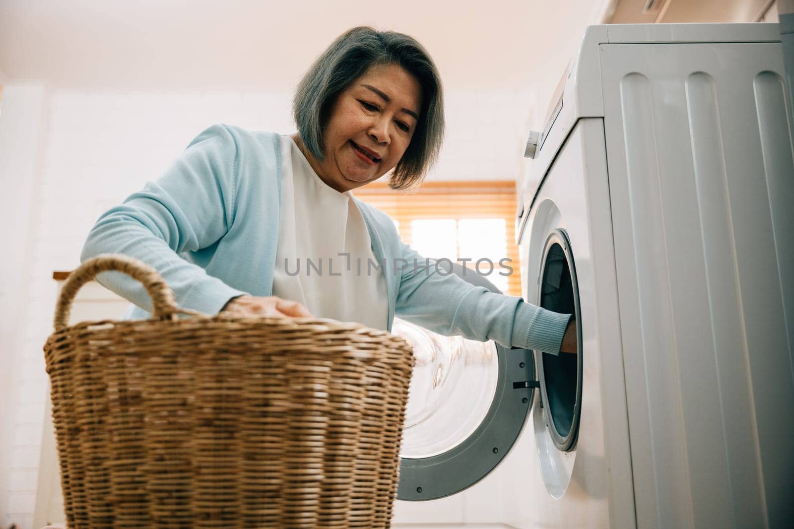 An old woman, a grandmother, loads laundry into the washing machine, smiling as she covers her routine. This portrait reflects a modern solution for family hygiene and happiness. by Sorapop
