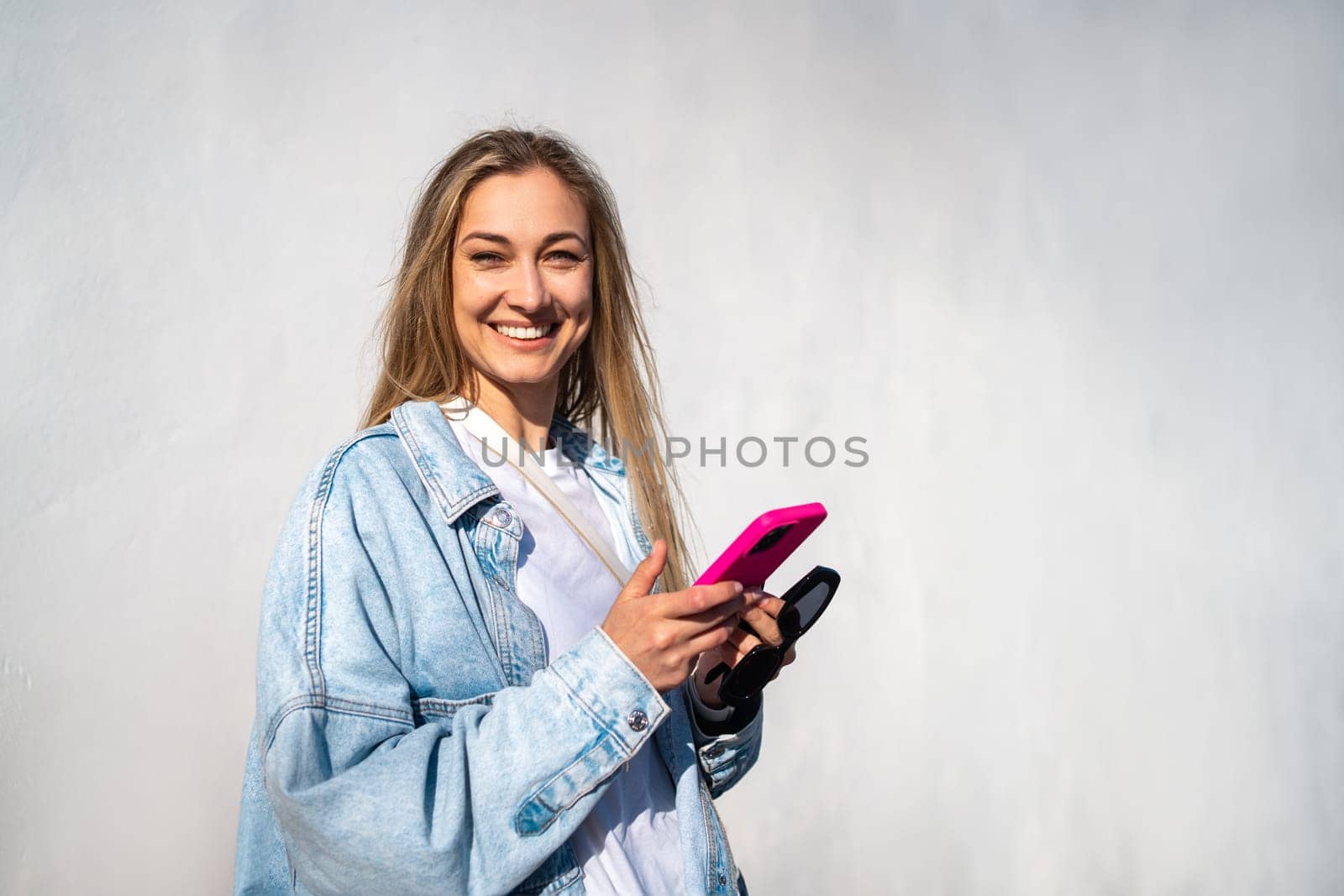 Portrait of cheerful young woman in denim shirt holding smartphone and looking at camera. Smiling female tourist standing against white wall on sunny day.