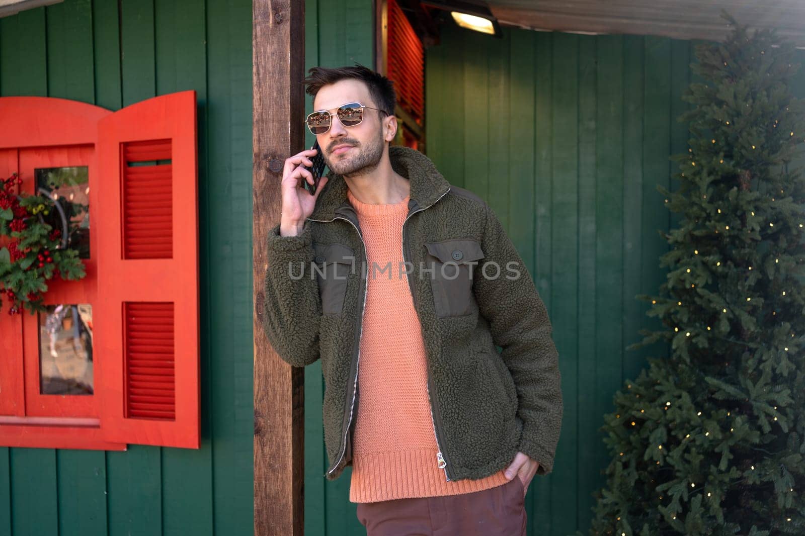 Caucasian man in sunglasses stands in festive town near Christmas tree, talking on smartphone. Stylish young guy in jacket extends holiday greetings through mobile device.