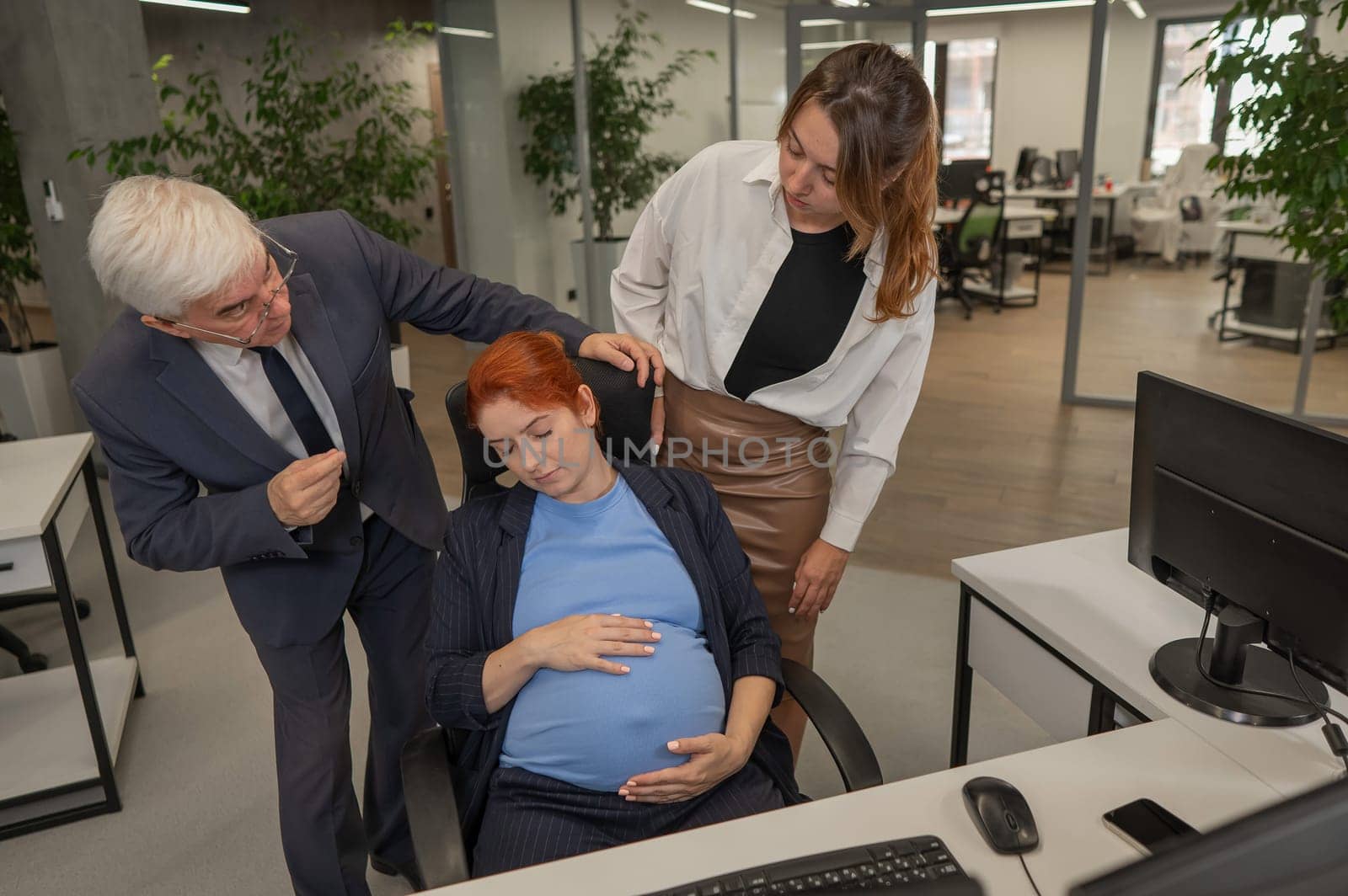 An elderly Caucasian man and woman look disapprovingly at their sleeping pregnant colleague in the office