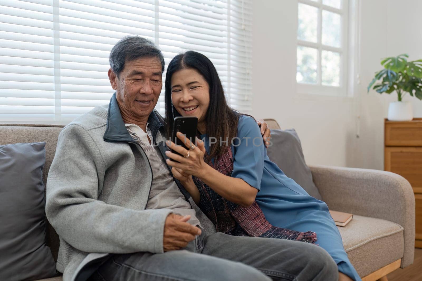 Happy married retirement couple looking at mobile using smartphone mobile technology device together, relaxing on couch doing online shopping.