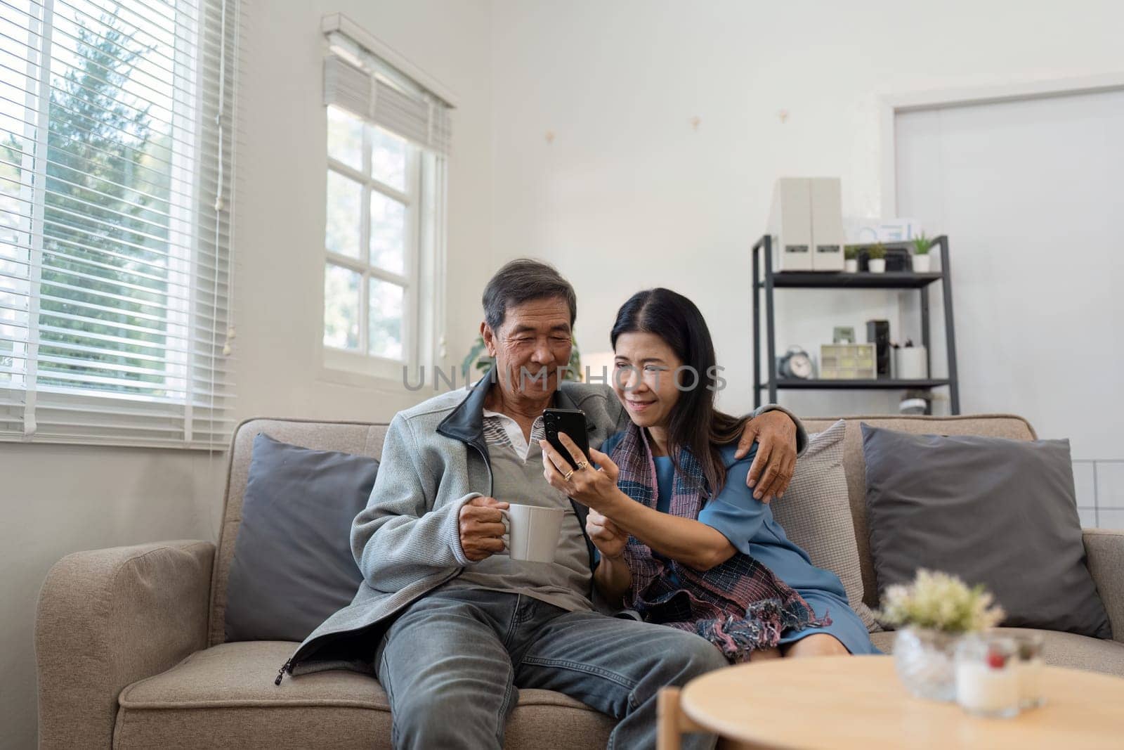 Happy married retirement couple looking at mobile using smartphone mobile technology device together, relaxing on couch doing online shopping.